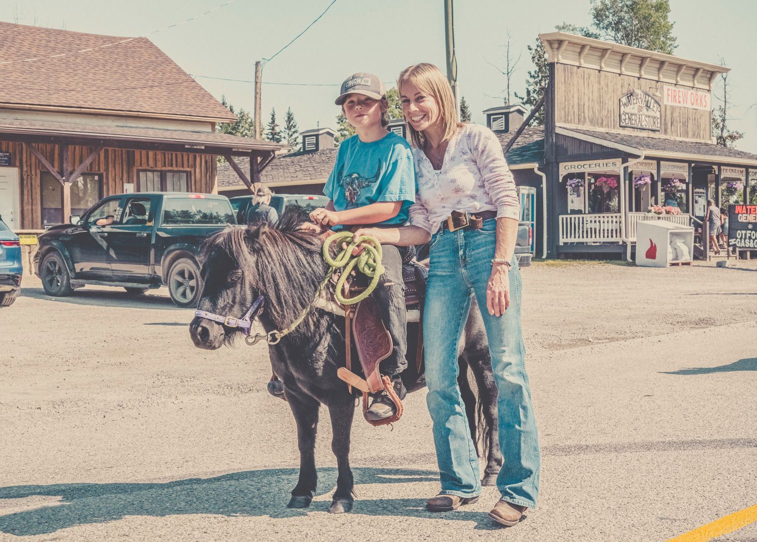 A person riding a small black pony, led by another individual, stands in front of rustic storefronts on a sunny day.