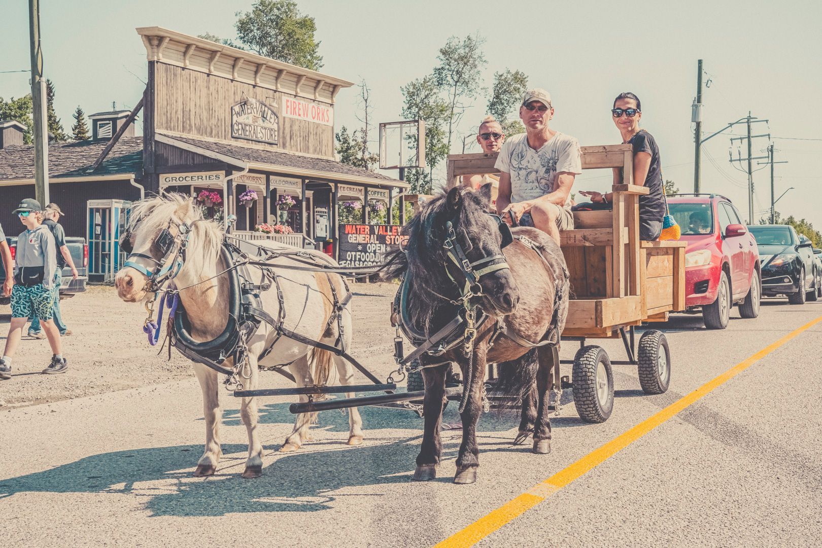 Two small horses pull a wooden carriage carrying three passengers past a rustic building and cars on a sunny day.