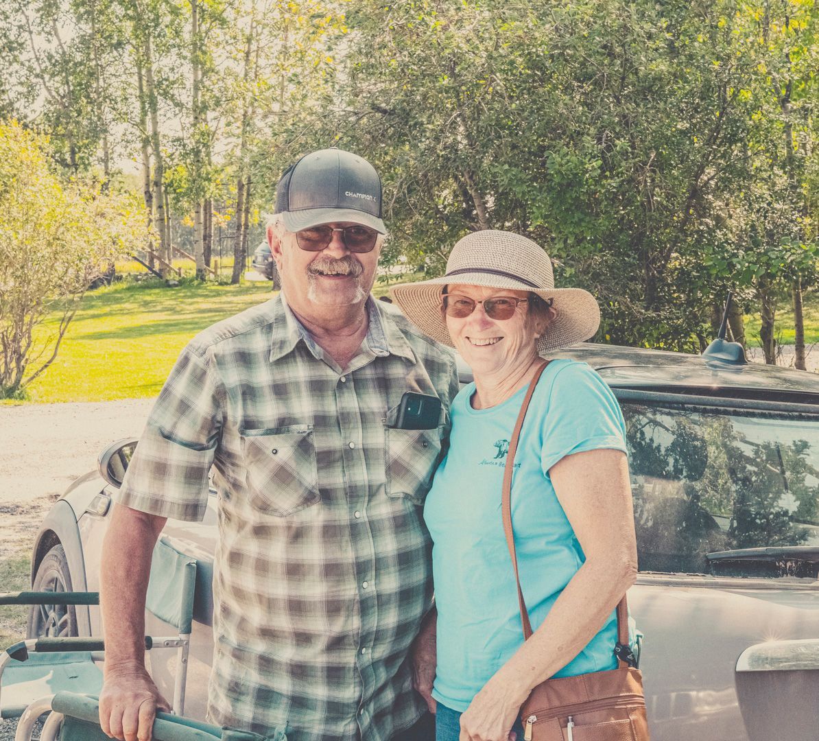 A smiling couple stands outdoors in front of a vehicle, wearing casual clothing, a plaid shirt, and a sun hat.