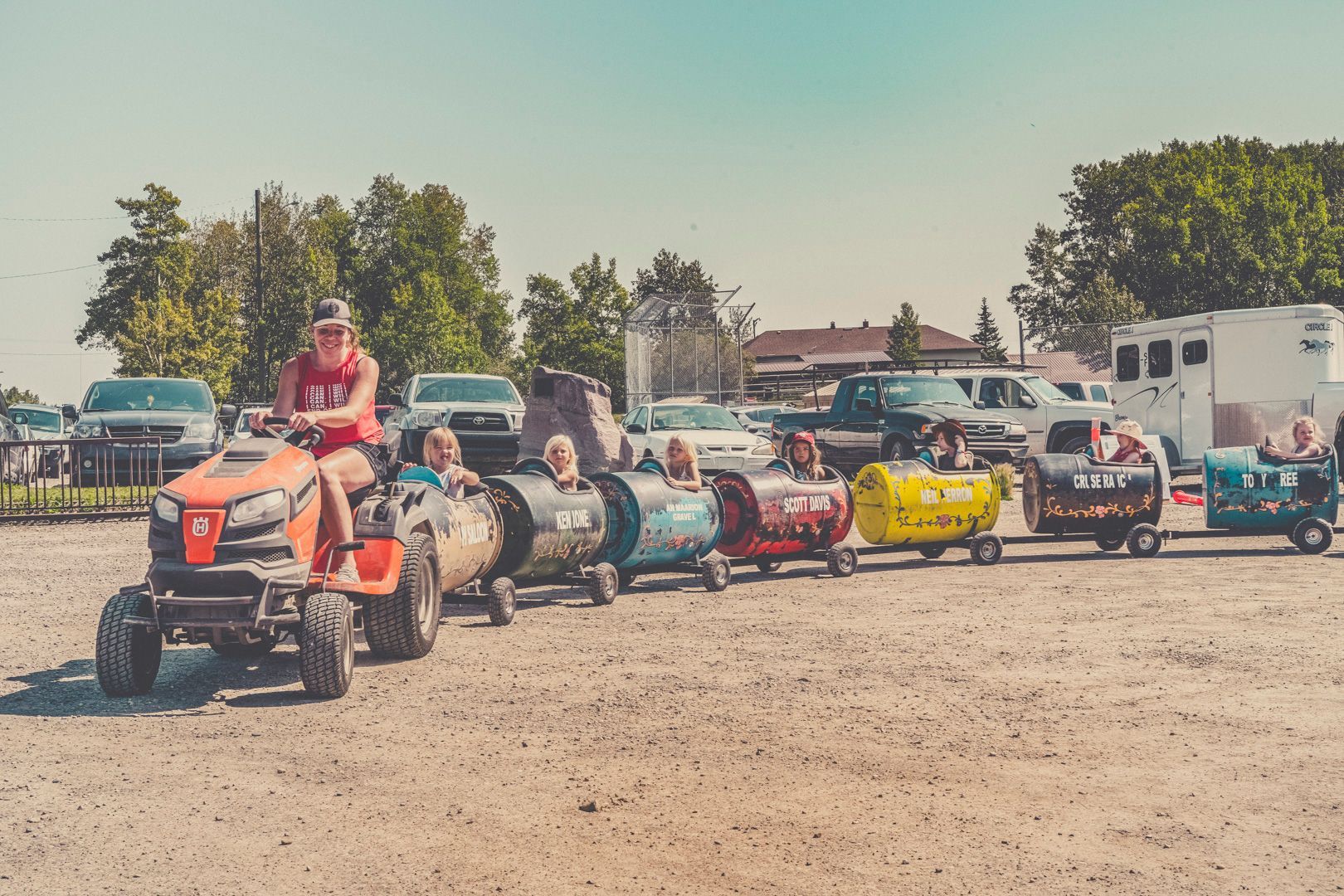 A person driving a lawn tractor pulling a train of colorful barrel cars carrying riders through an outdoor parking area.