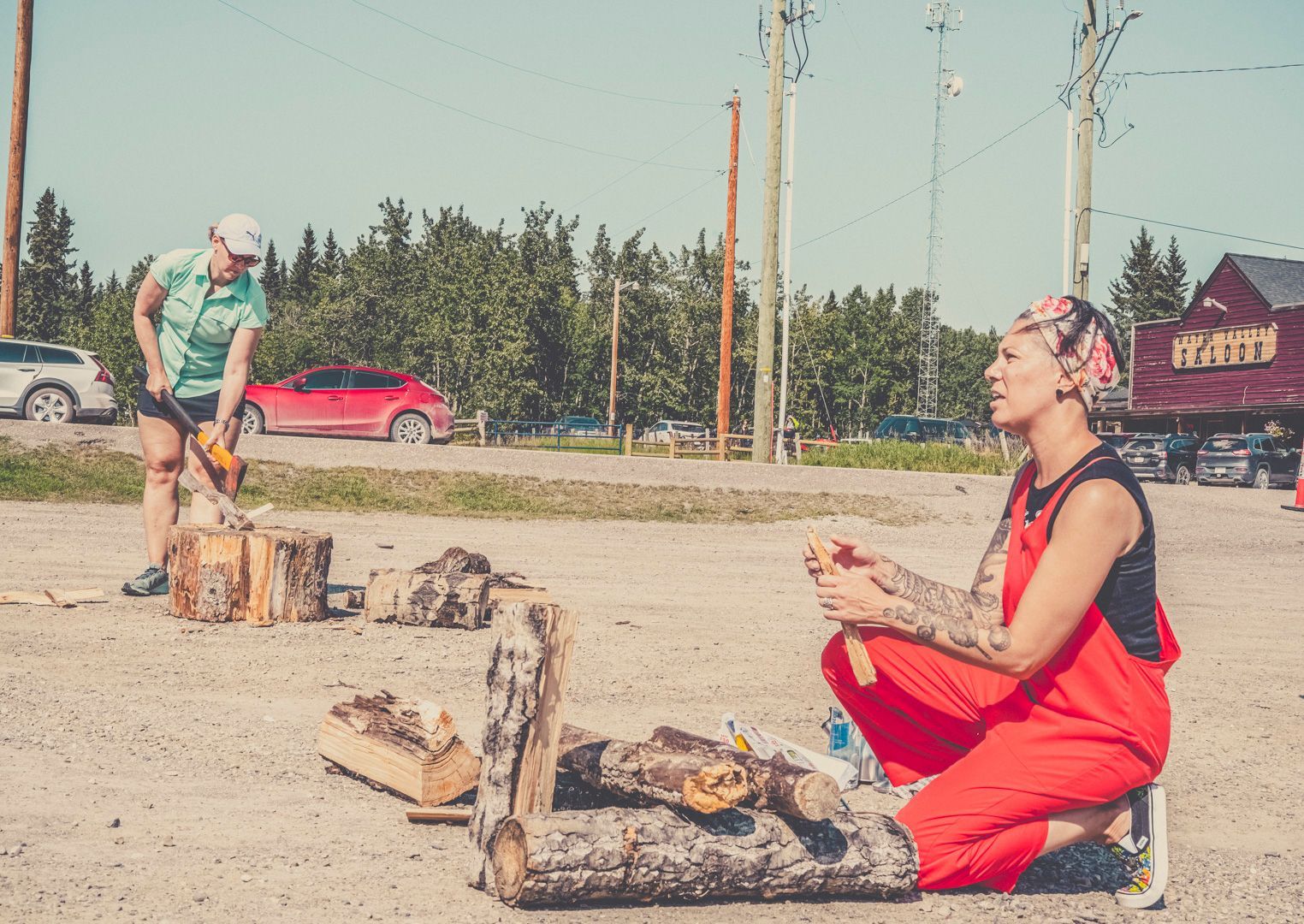 Two people prepare firewood outdoors on a gravel lot near trees, a red building, and parked cars.