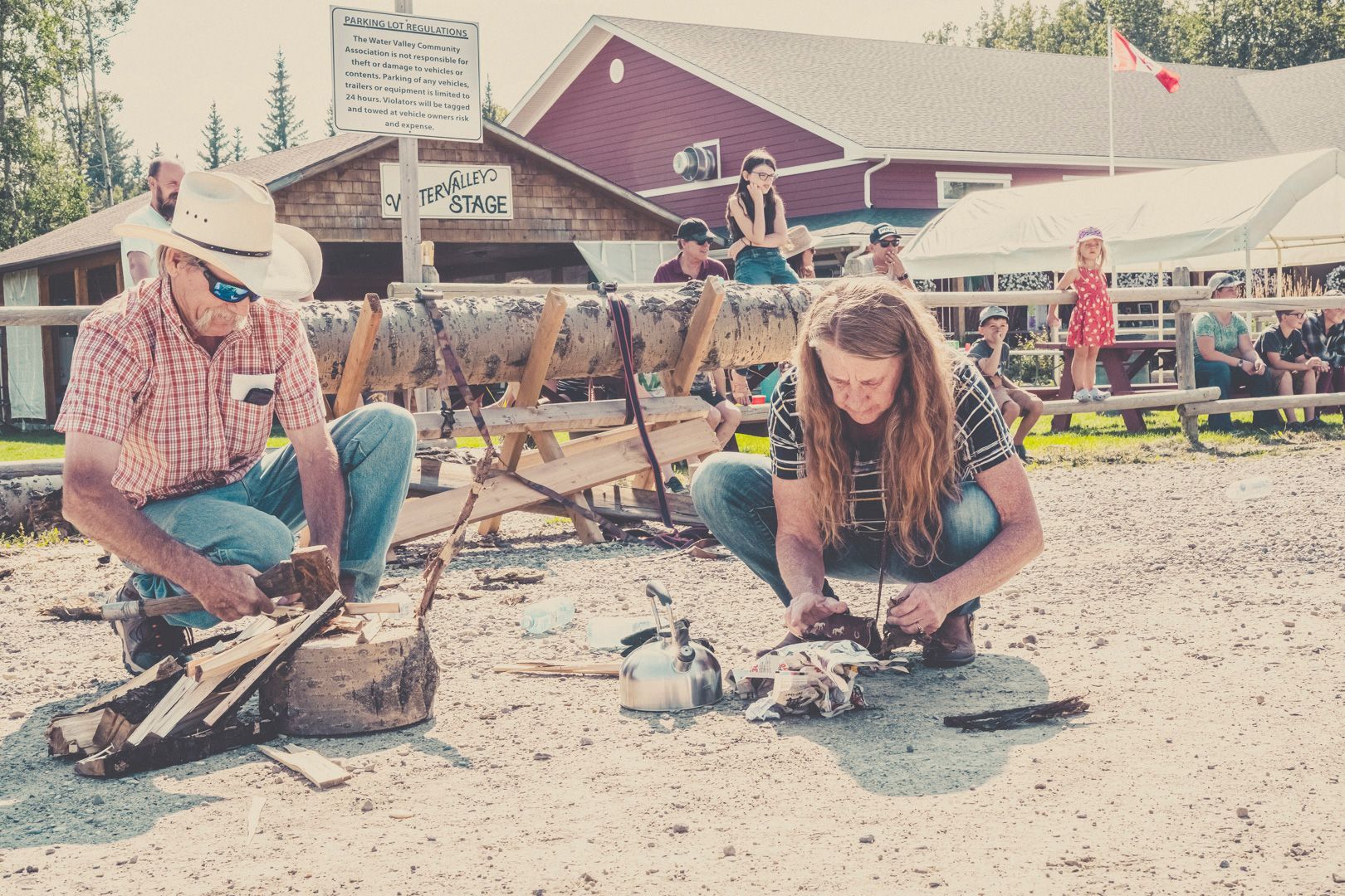 Two people demonstrate traditional woodcraft, one using a mallet and chisel, in a sunlit, outdoor fair setting.