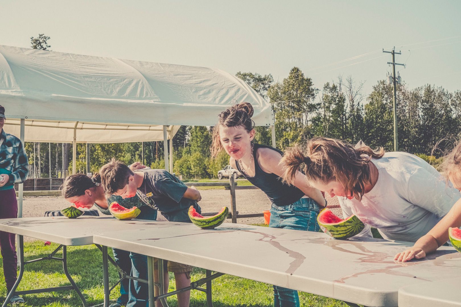 Participants lean over a long table outdoors, competing in a watermelon-eating contest.