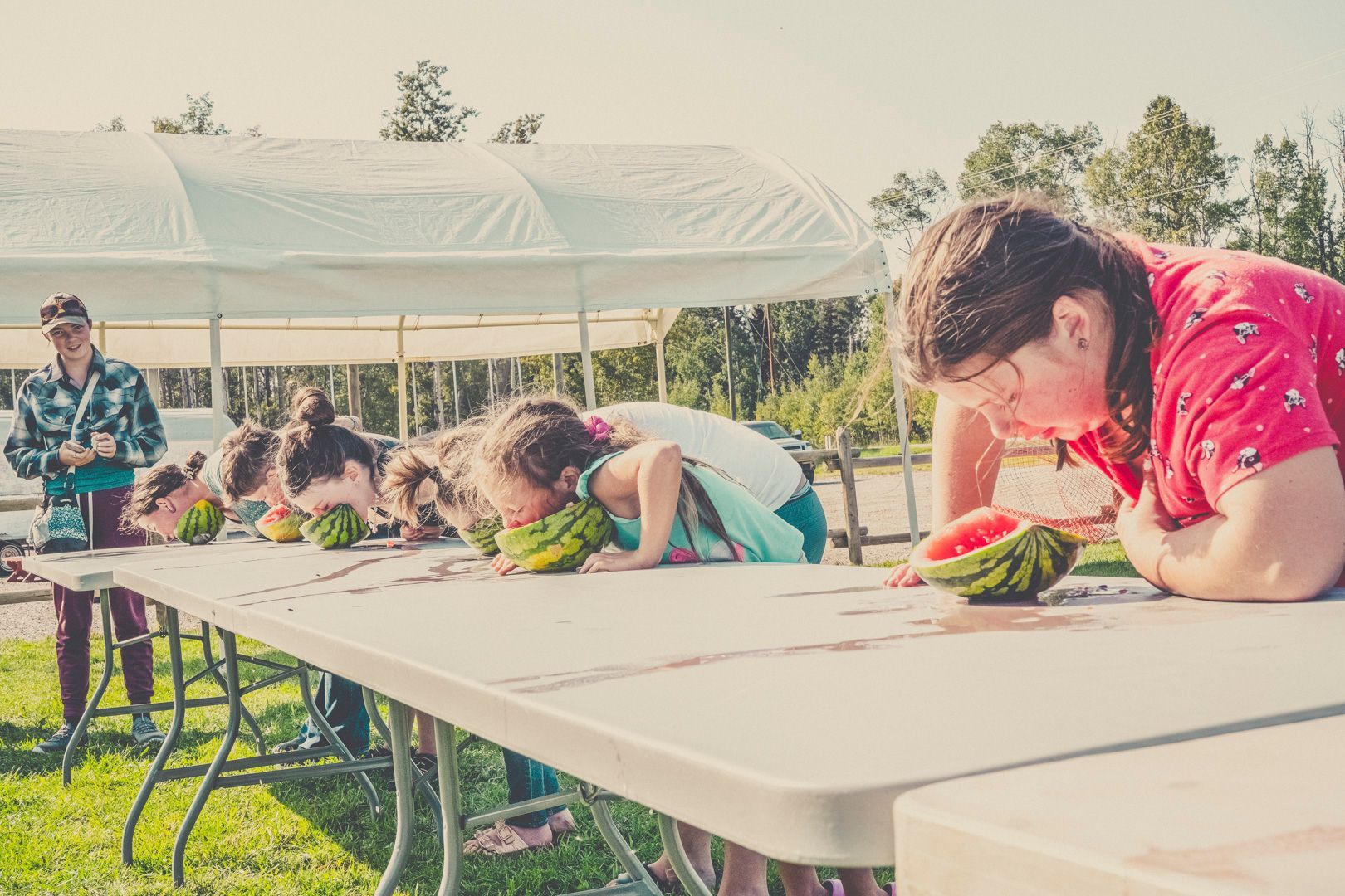 A group of people lean over a long white table outdoors, eating slices of watermelon without using their hands.