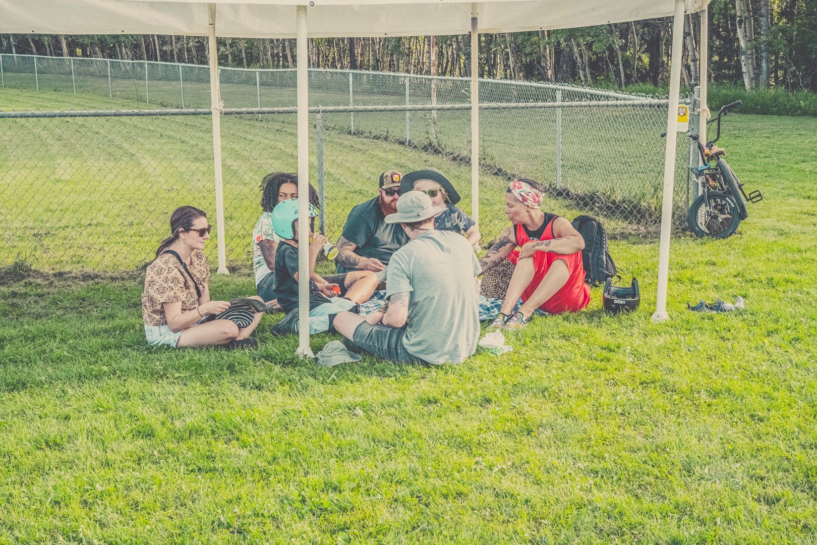 A group of people sit together under a white canopy tent in a grassy field near a chain-link fence.