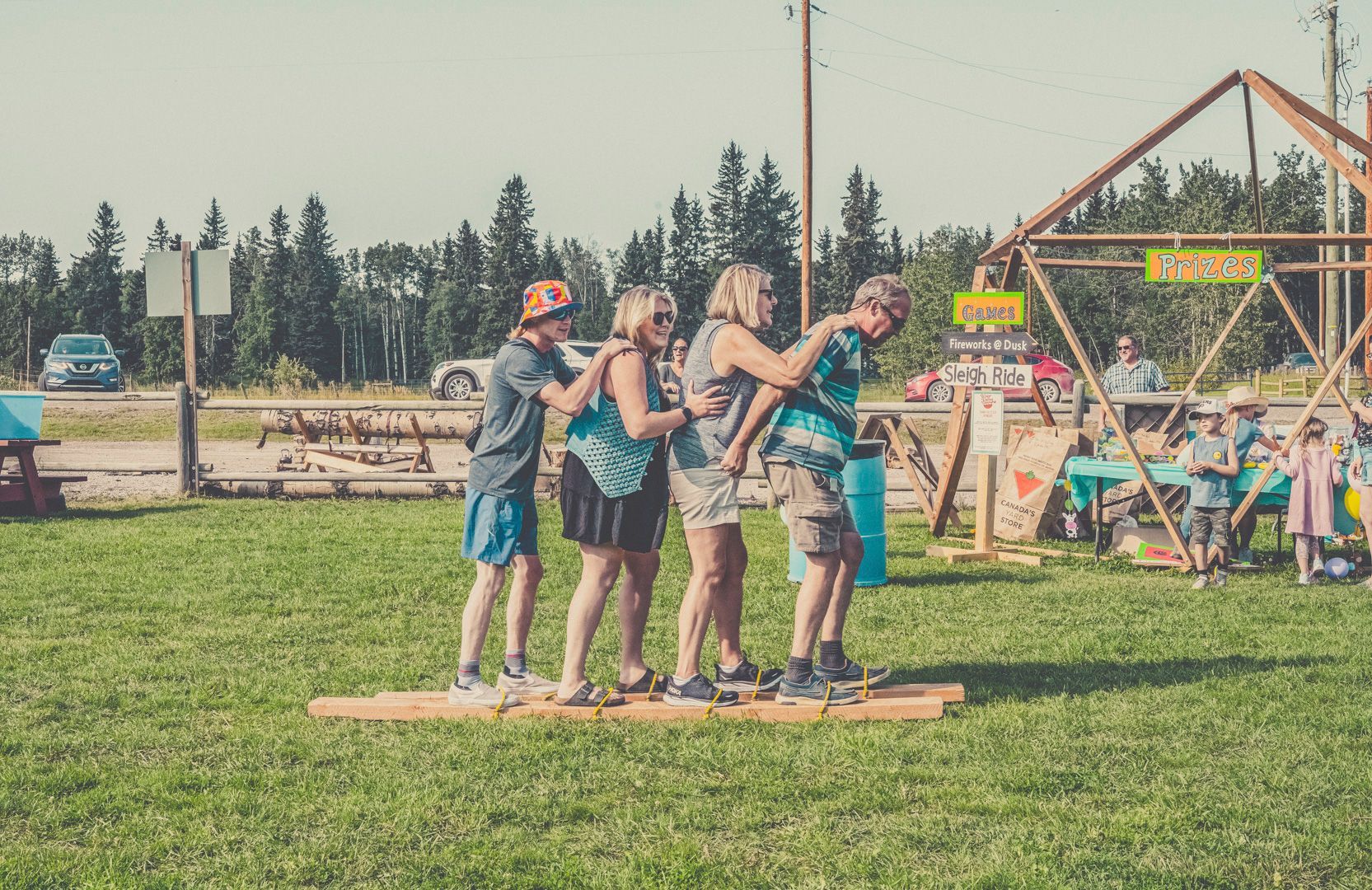 Four people walk in unison on a long wooden plank at an outdoor grassy festival.