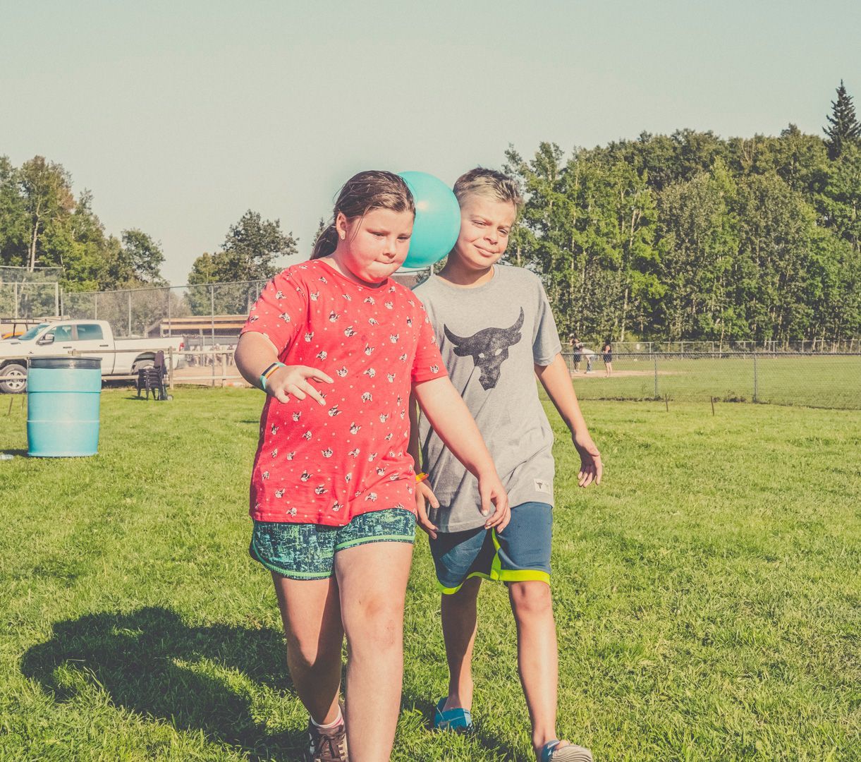Two young people walk across a grassy field outdoors, one holding a blue balloon behind them.