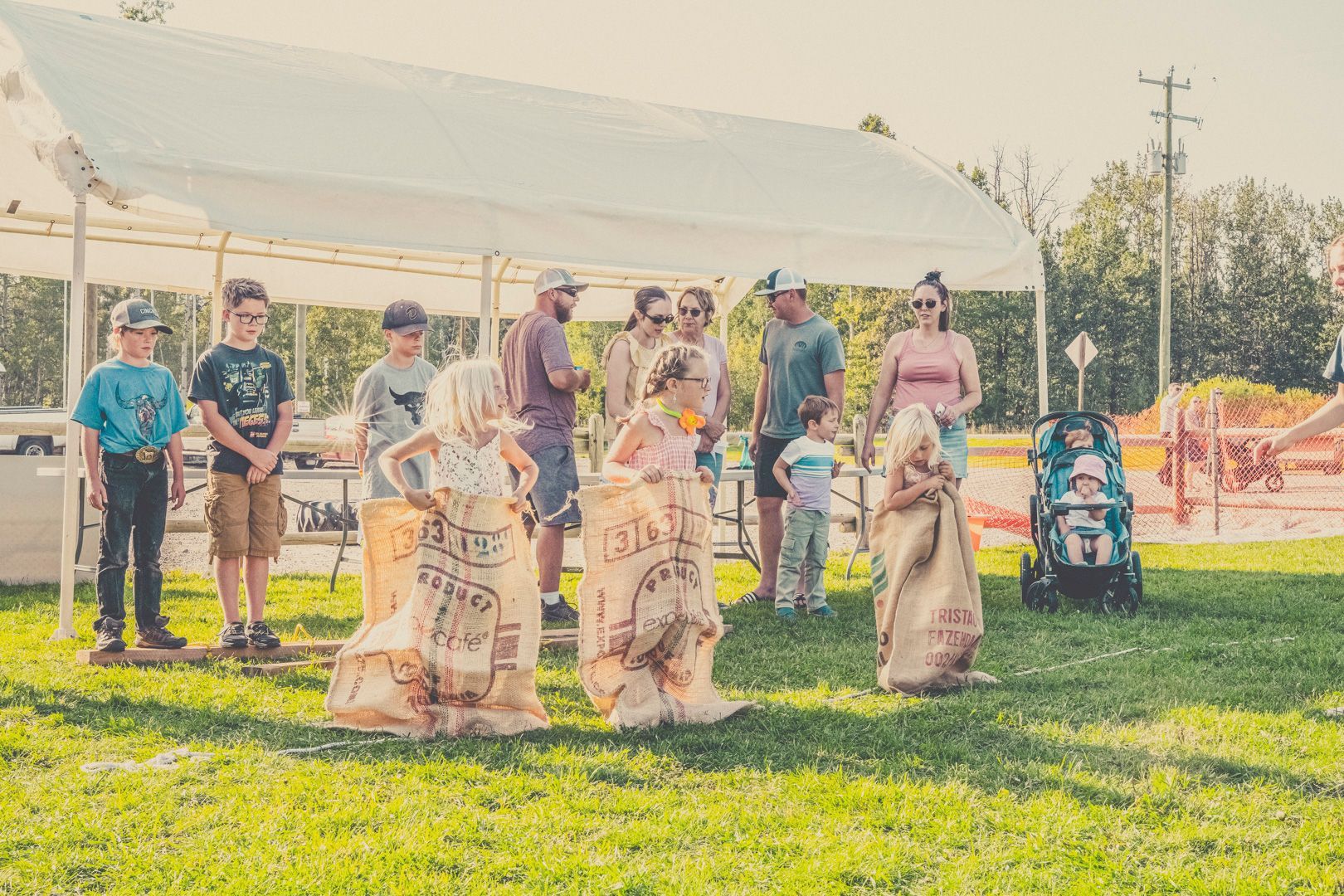 Children in burlap sacks participate in a potato sack race on a grassy field under a white event tent.