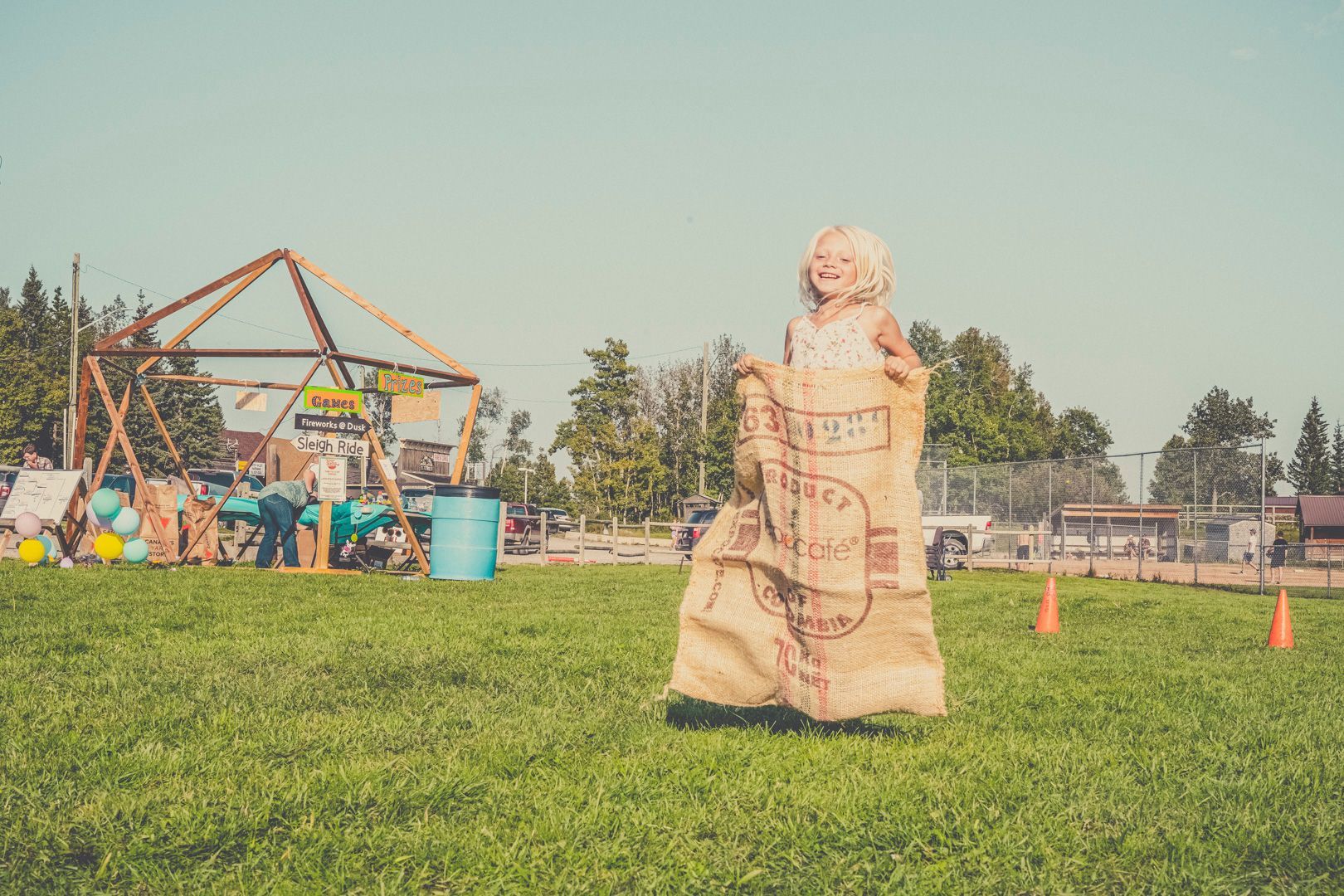 A person laughing while jumping in a burlap sack race on a grassy field with a wooden structure in the background.