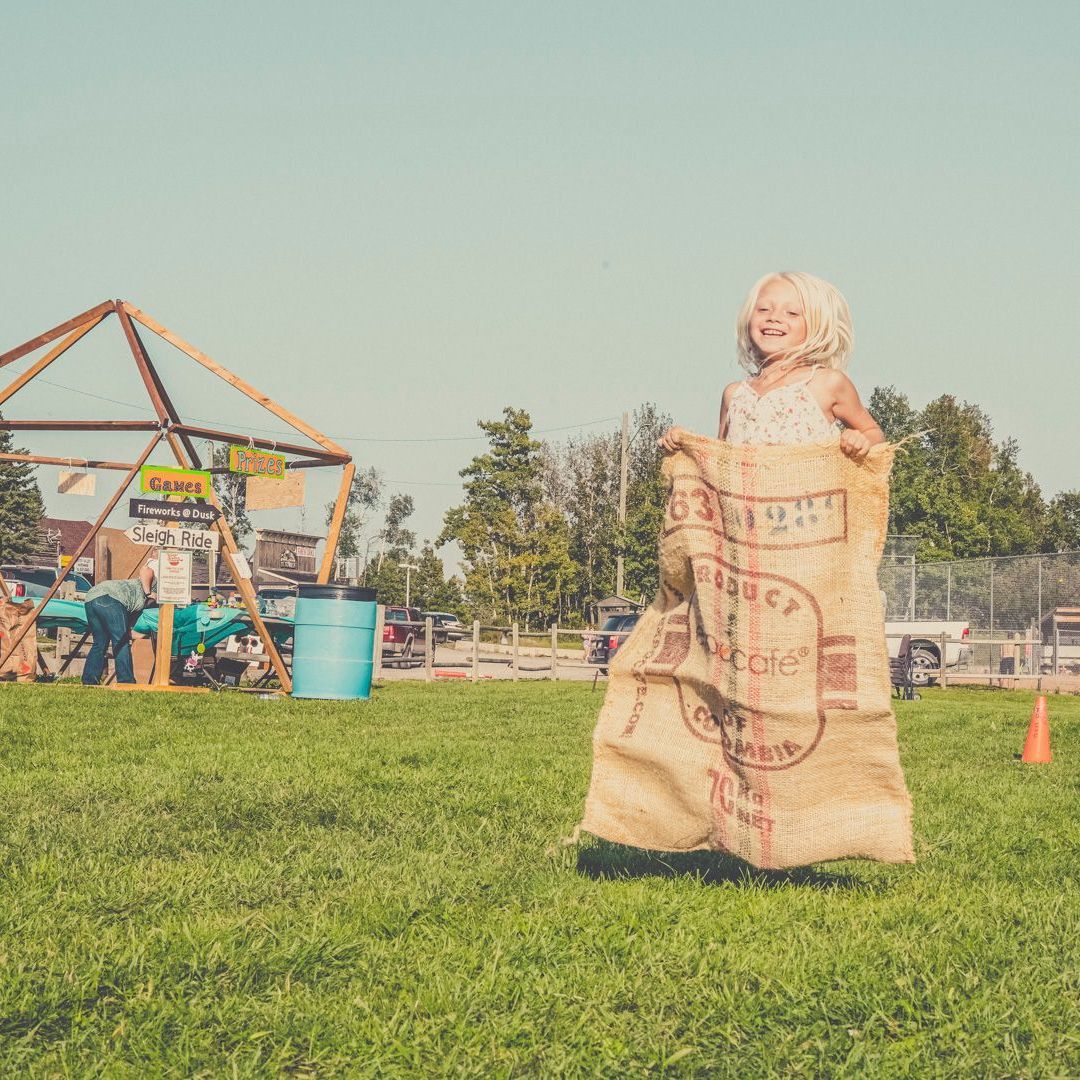 A smiling person jumps inside a tan burlap sack during a race on a grassy field near a wooden frame structure.