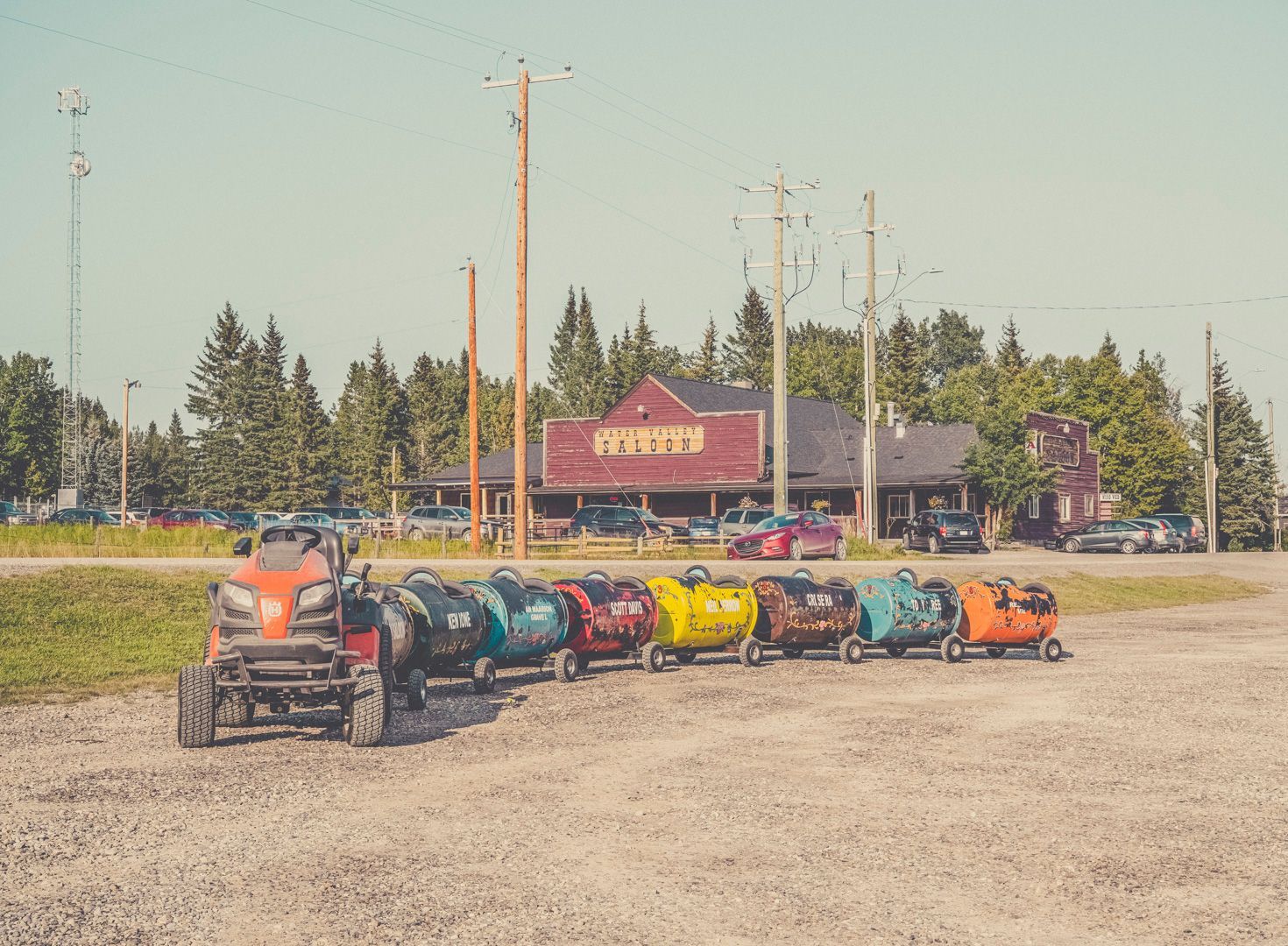 A riding lawn mower pulling a train of small, brightly colored trailers across a gravel lot in front of a store.