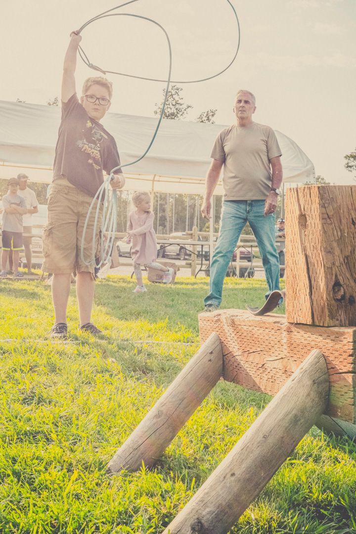 A young person practices lassoing a wooden steer target in a grassy field while an adult watches.