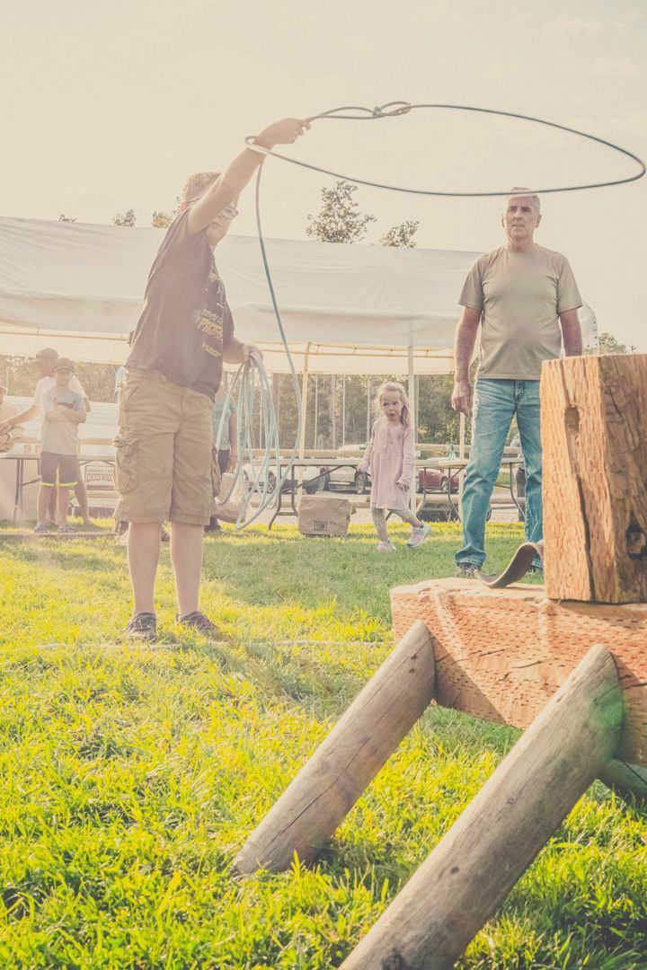 A person throws a lasso towards a wooden stump in a grassy field with spectators nearby.