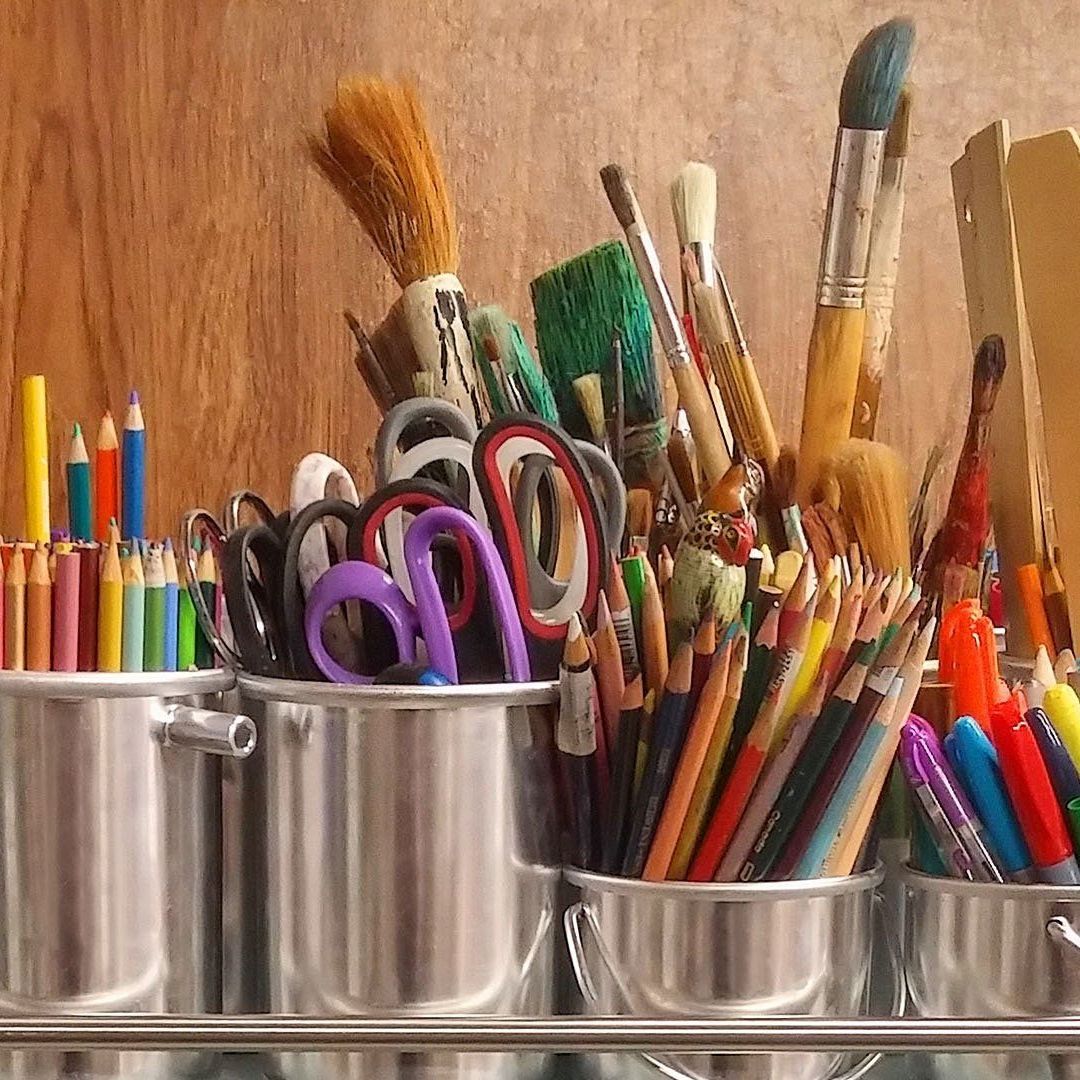 Four metal cups holding a variety of colorful pencils, paintbrushes, and pairs of scissors against a wooden background.