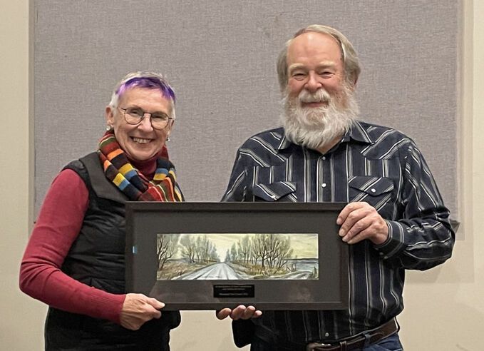 Two people stand side-by-side, smiling, while holding a framed panoramic landscape print in front of a gray wall.