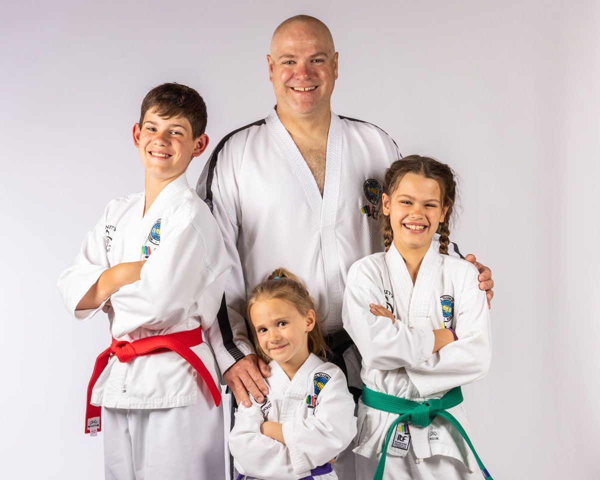 A smiling martial arts instructor and three students in white uniforms with colorful belts pose together against a white.