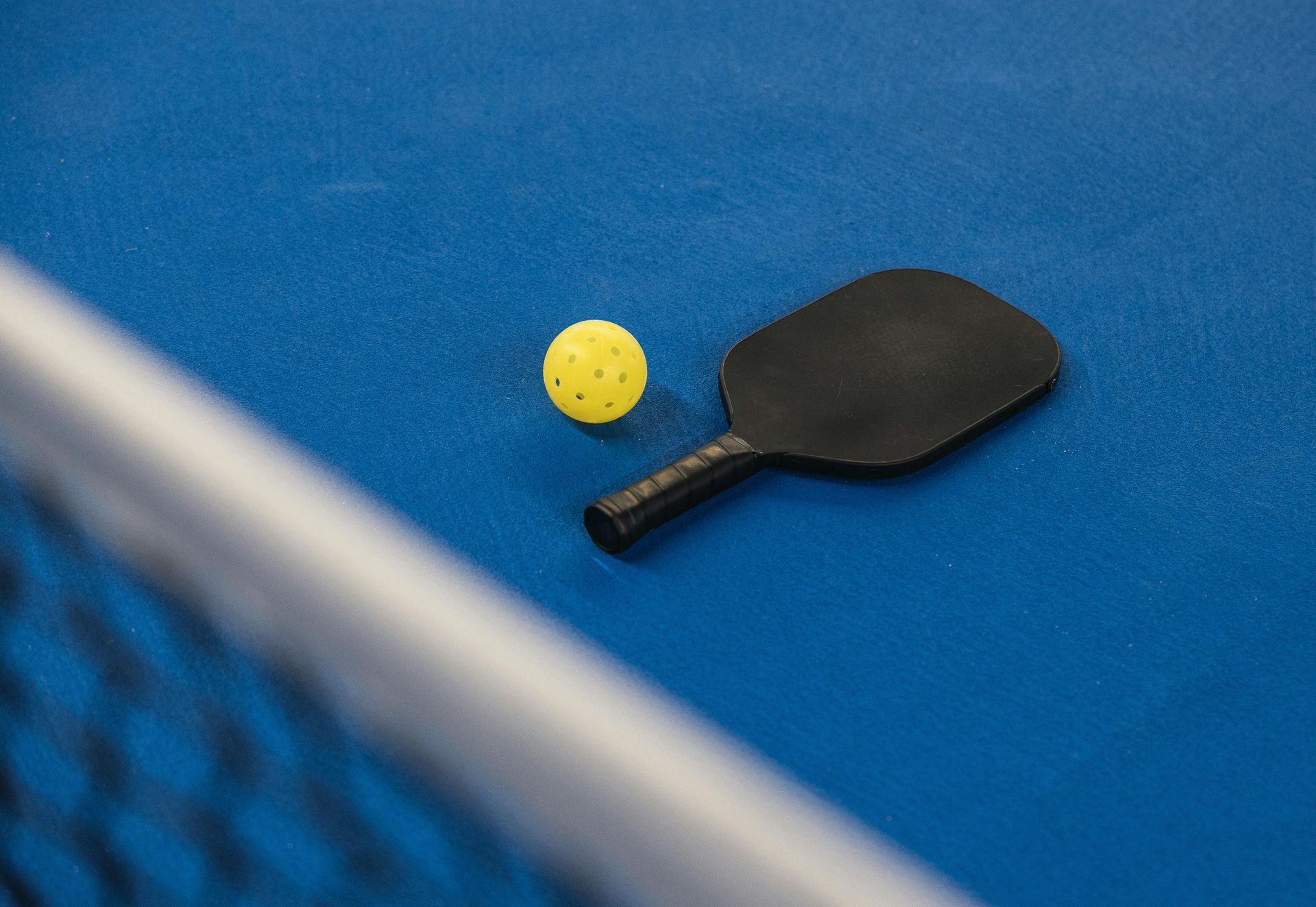 A yellow pickleball and a black paddle sit on a bright blue court near a blurred net.