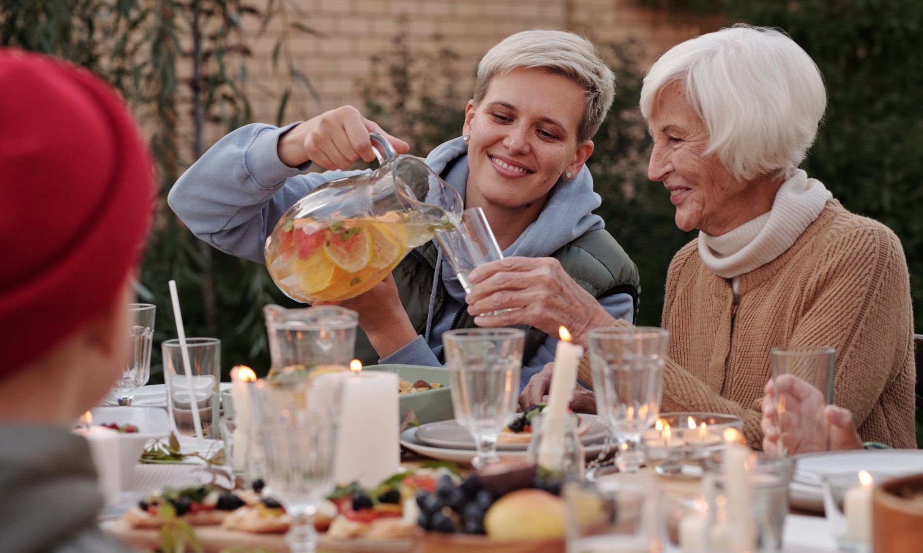 People gather for an outdoor meal as one person pours a glass of fruit-infused beverage at a table with lit candles.