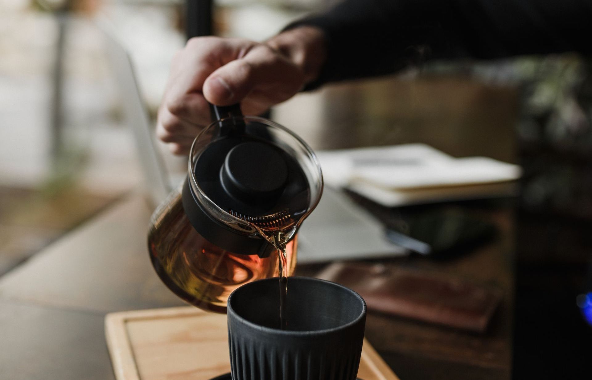 A person pours hot coffee from a clear glass carafe into a dark, ribbed mug at a desk with a laptop and notebook nearby.
