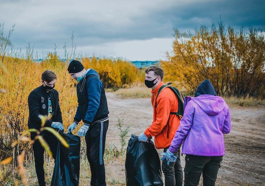 Four people wearing masks and casual clothing collect litter in black bags outdoors among yellow autumnal trees.