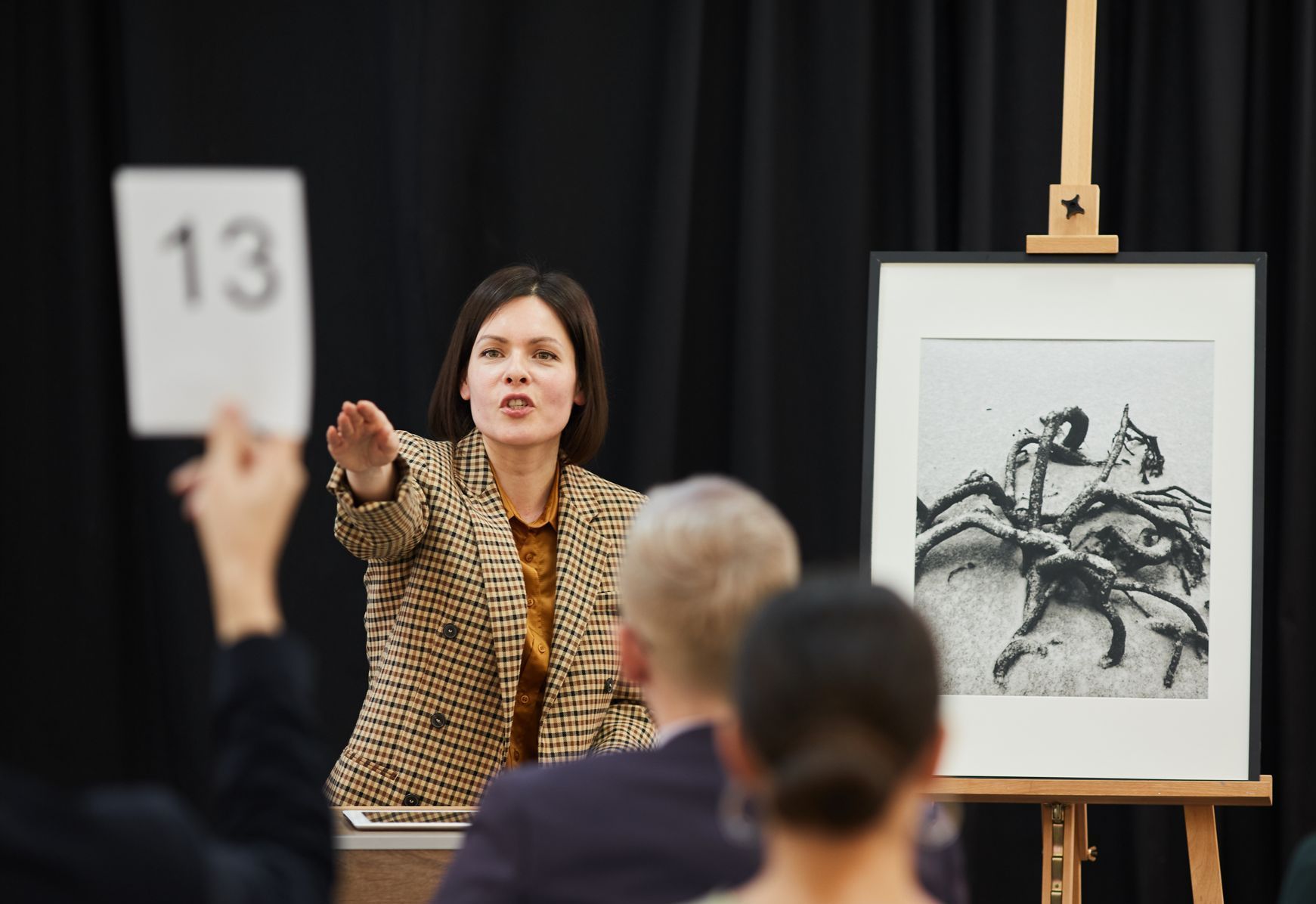 An auctioneer points to a bidder holding a paddle with the number 13, with a framed artwork on an easel nearby.