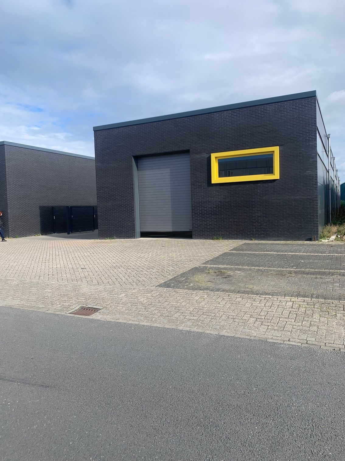Een modern, donkergrijs industrieel gebouw met een grote roldeur en een enkel horizontaal raam met een felgeel kozijn.
