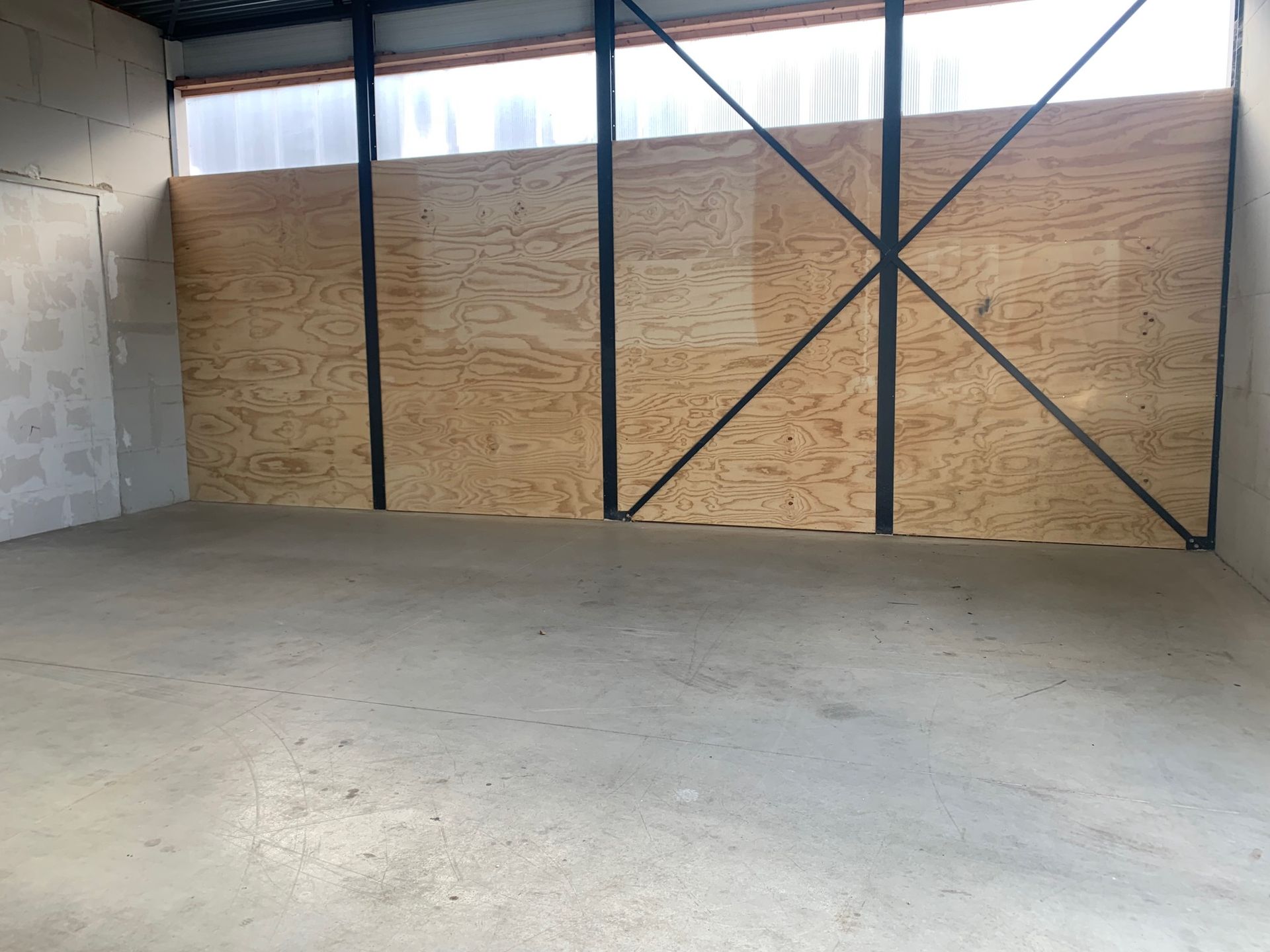 Een ruim industrieel interieur met betonnen vloeren, witte bakstenen muren en grote multiplex panelen omlijst door zwarte balken.