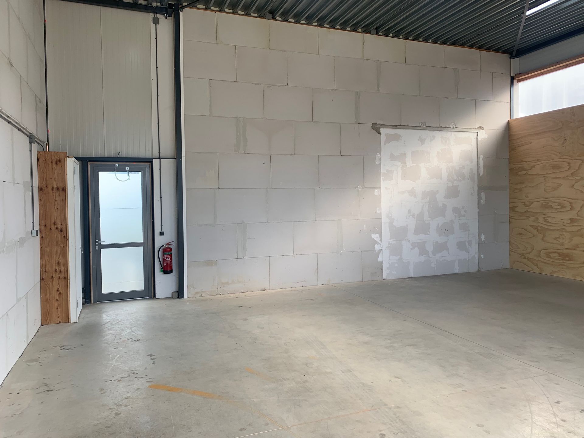 Een interieurfoto van een magazijn met grijze betonnen muren, een enkele glazen deur en multiplex panelen aan de rechterkant.