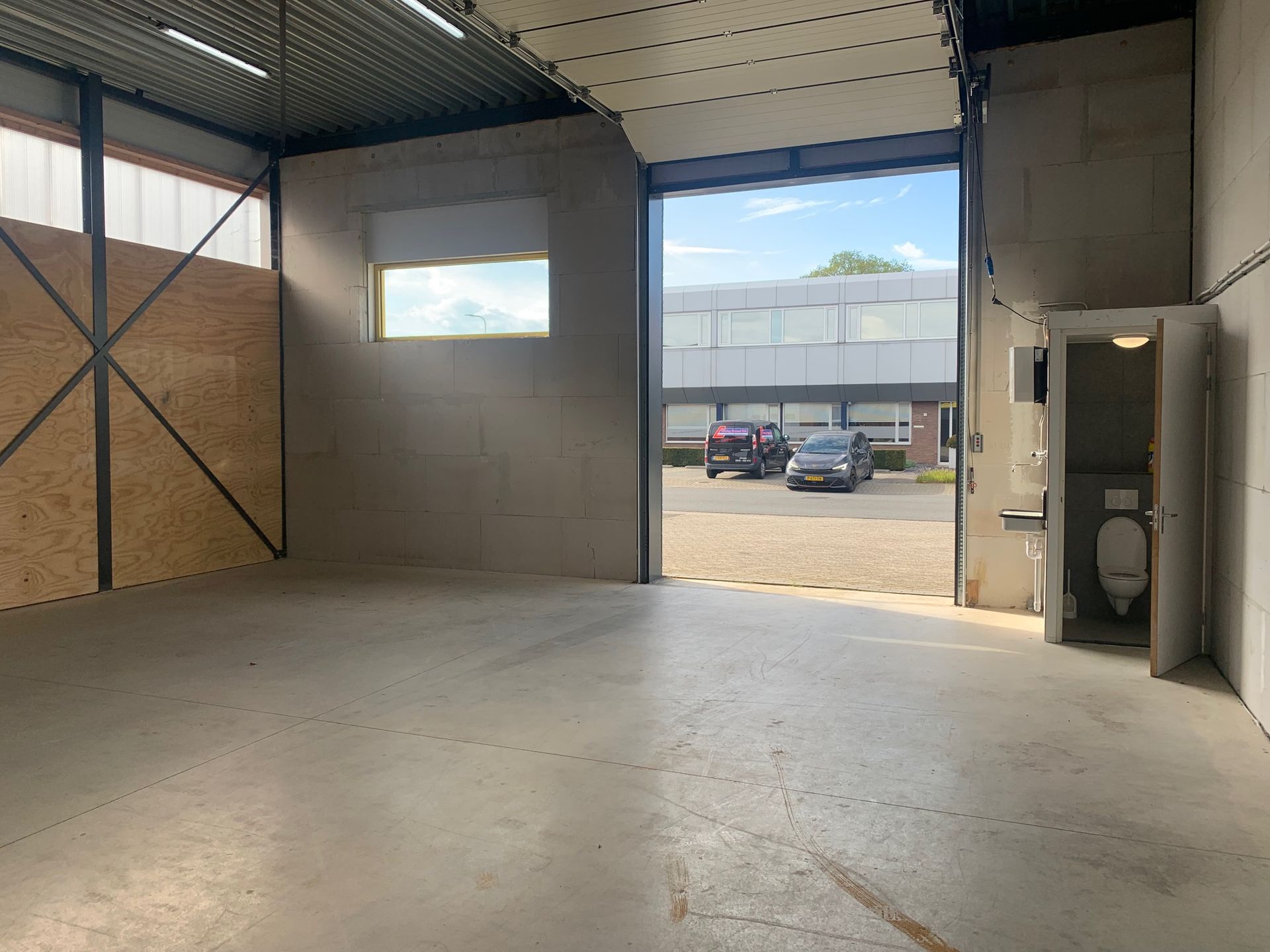 Een open magazijninterieur met betonnen muren, een grote roldeur naar een parkeerplaats en een klein toilet in de hoek.