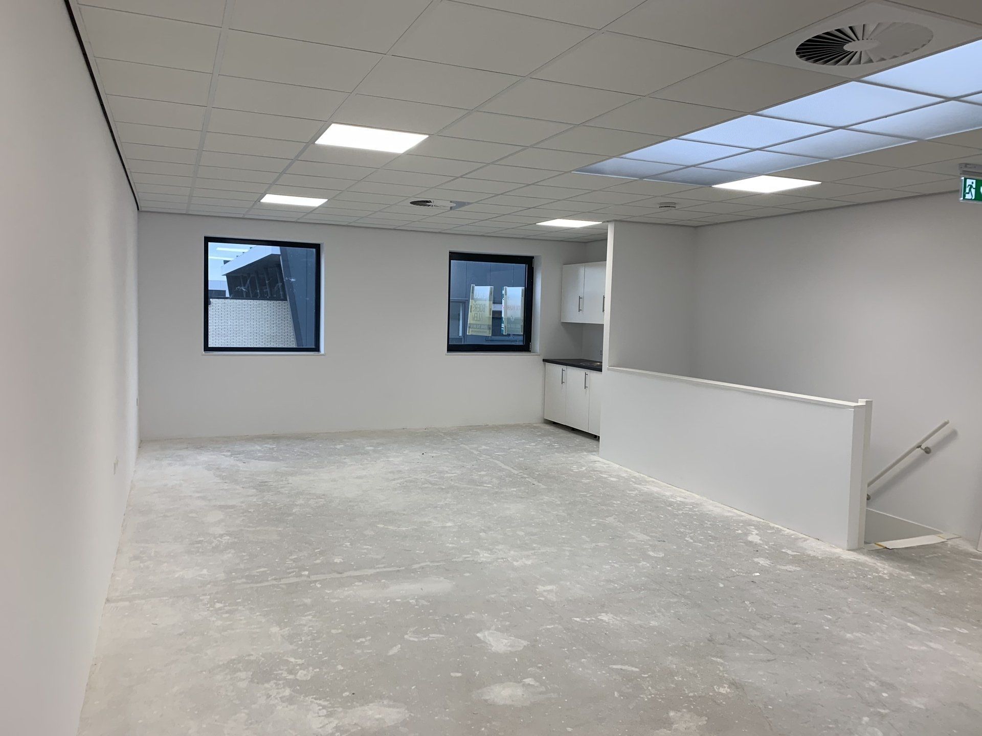 Een lege kantoorruimte met witte muren, een grijze betonnen vloer, twee ramen, inbouwspots in het plafond en een wit aanrechtblad.
