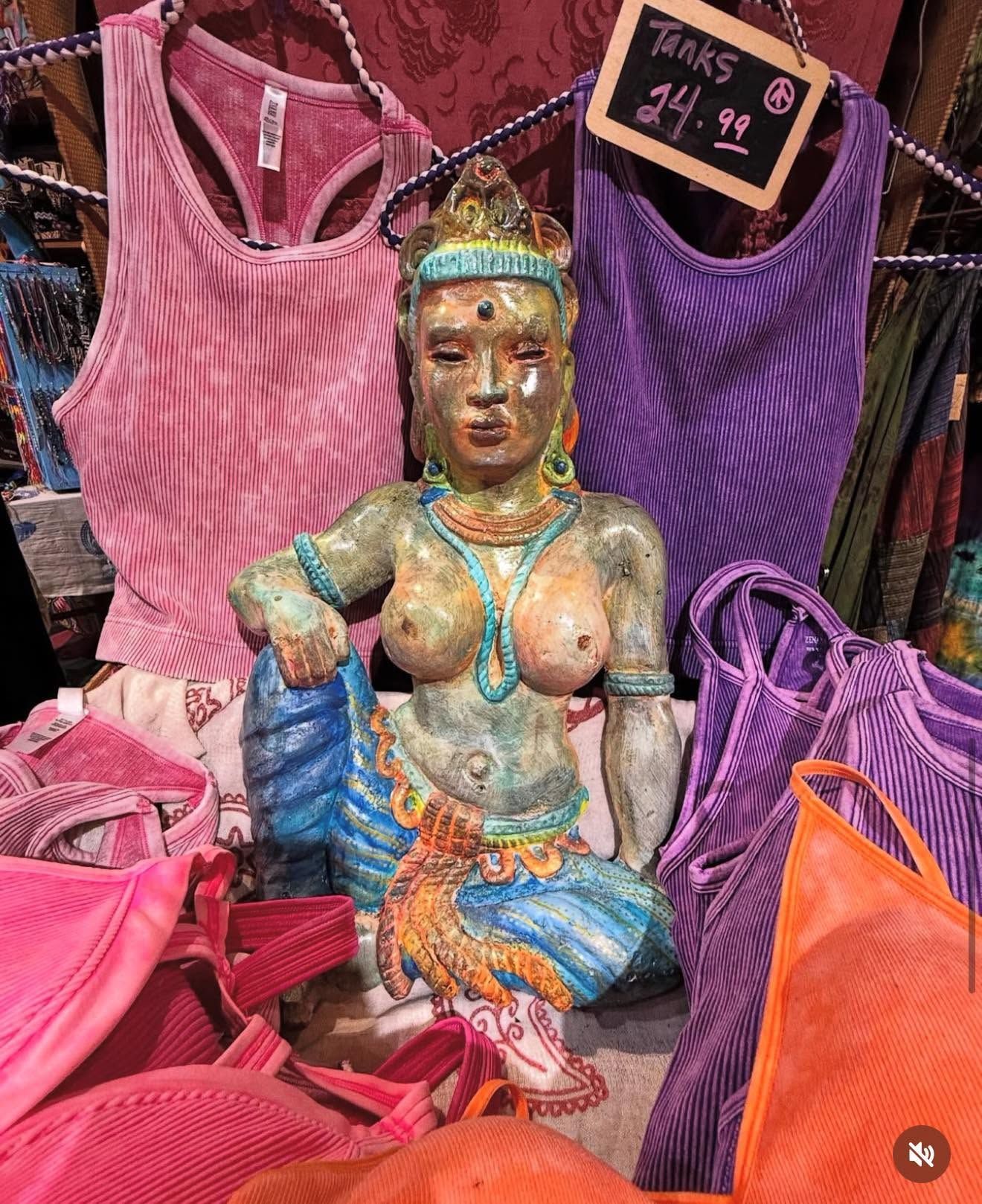 Colorful market stall with a gold-painted seated statue surrounded by pink, purple, and orange knit tops and bags