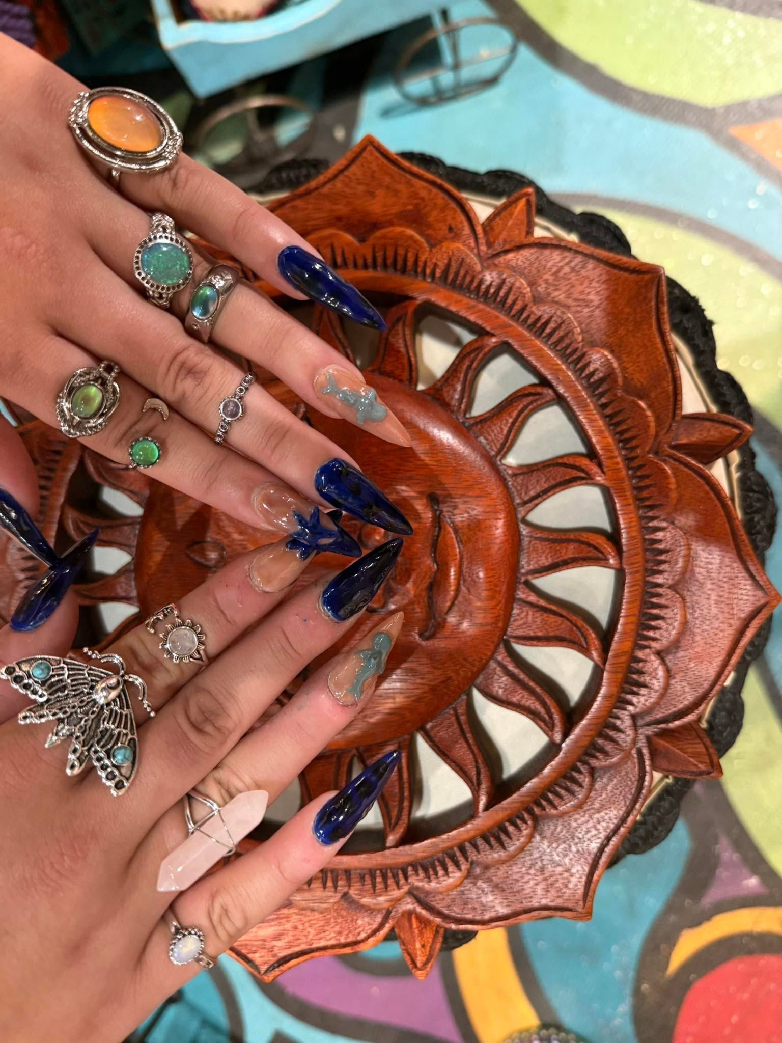 Hands with jeweled, long blue nails arranged around a carved wooden decorative bowl.