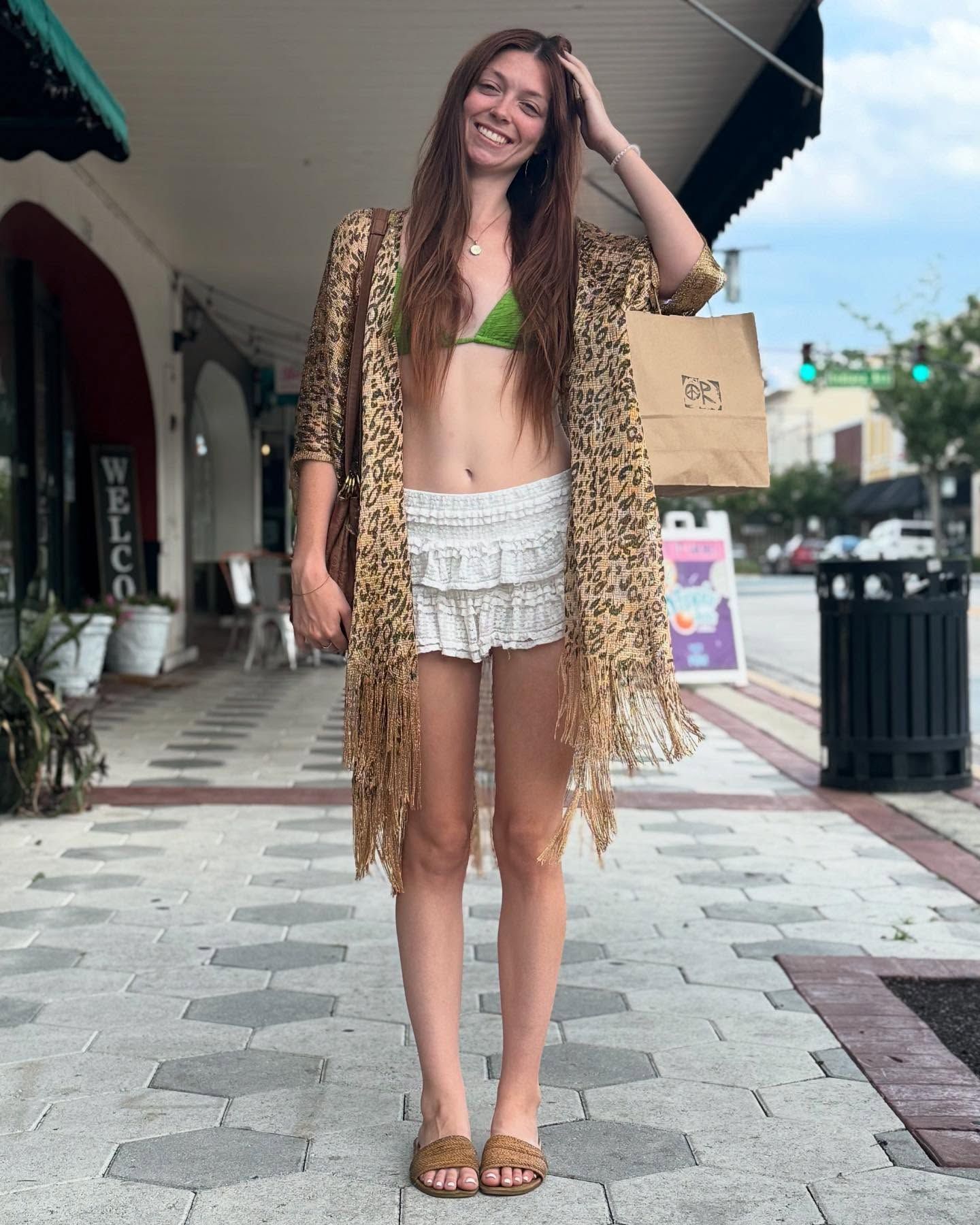 Woman smiling in beachwear and a patterned cover-up, standing on a sidewalk with shopping bags.