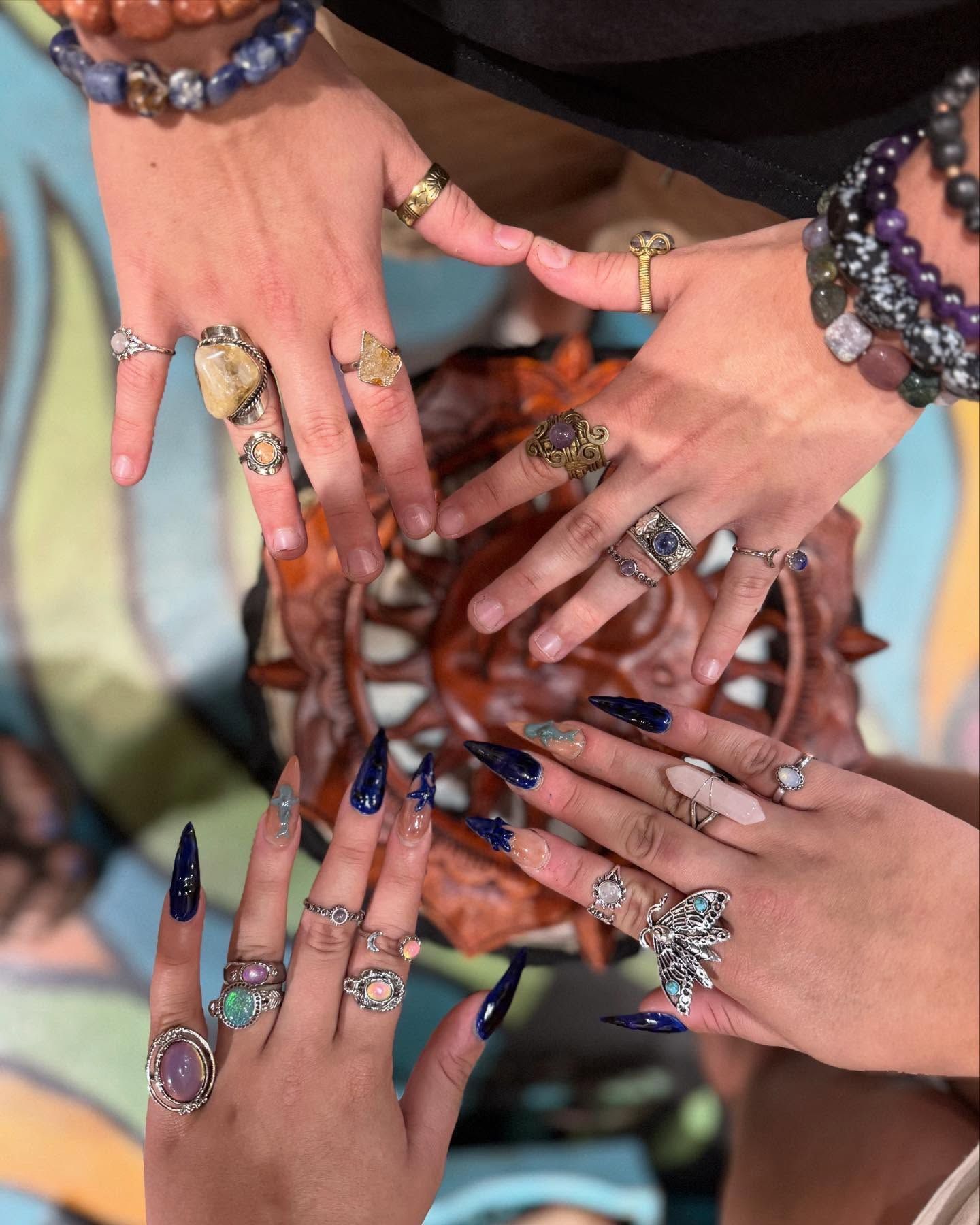Many hands with colorful nail art and rings stacked in a circle, showing bracelets and jewelry