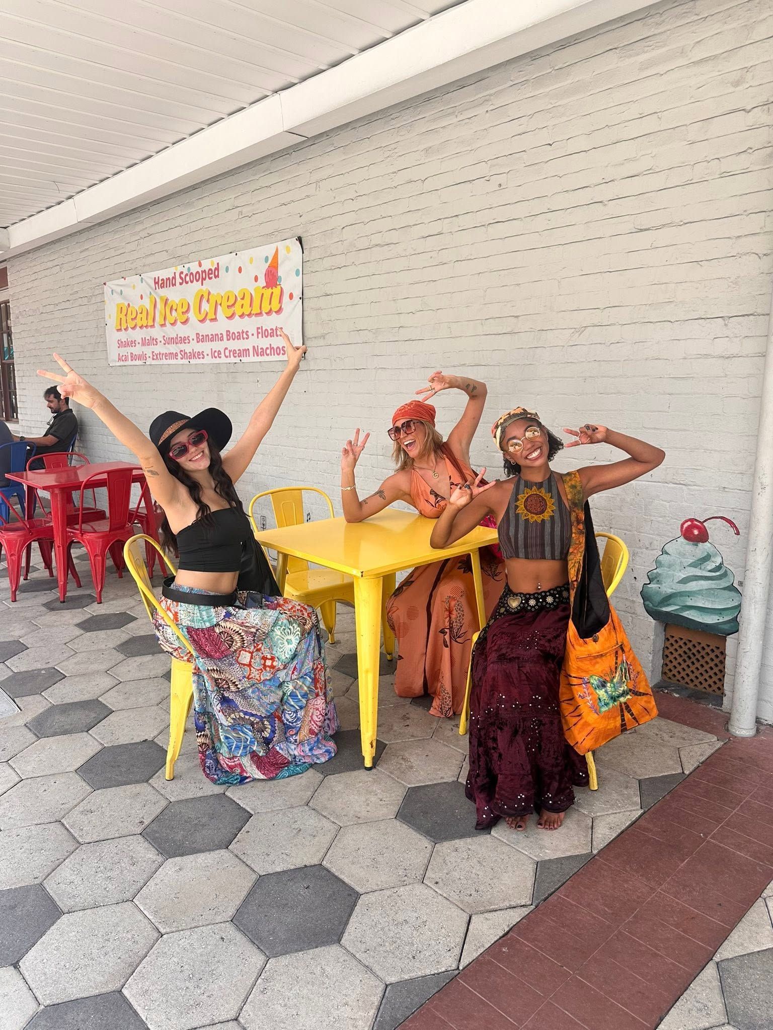 Three performers in colorful costumes pose dancing outside a café with yellow tables and chairs
