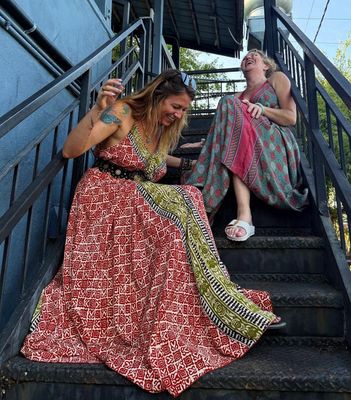 Two women in colorful dresses sit laughing on outdoor stairs under a metal staircase.