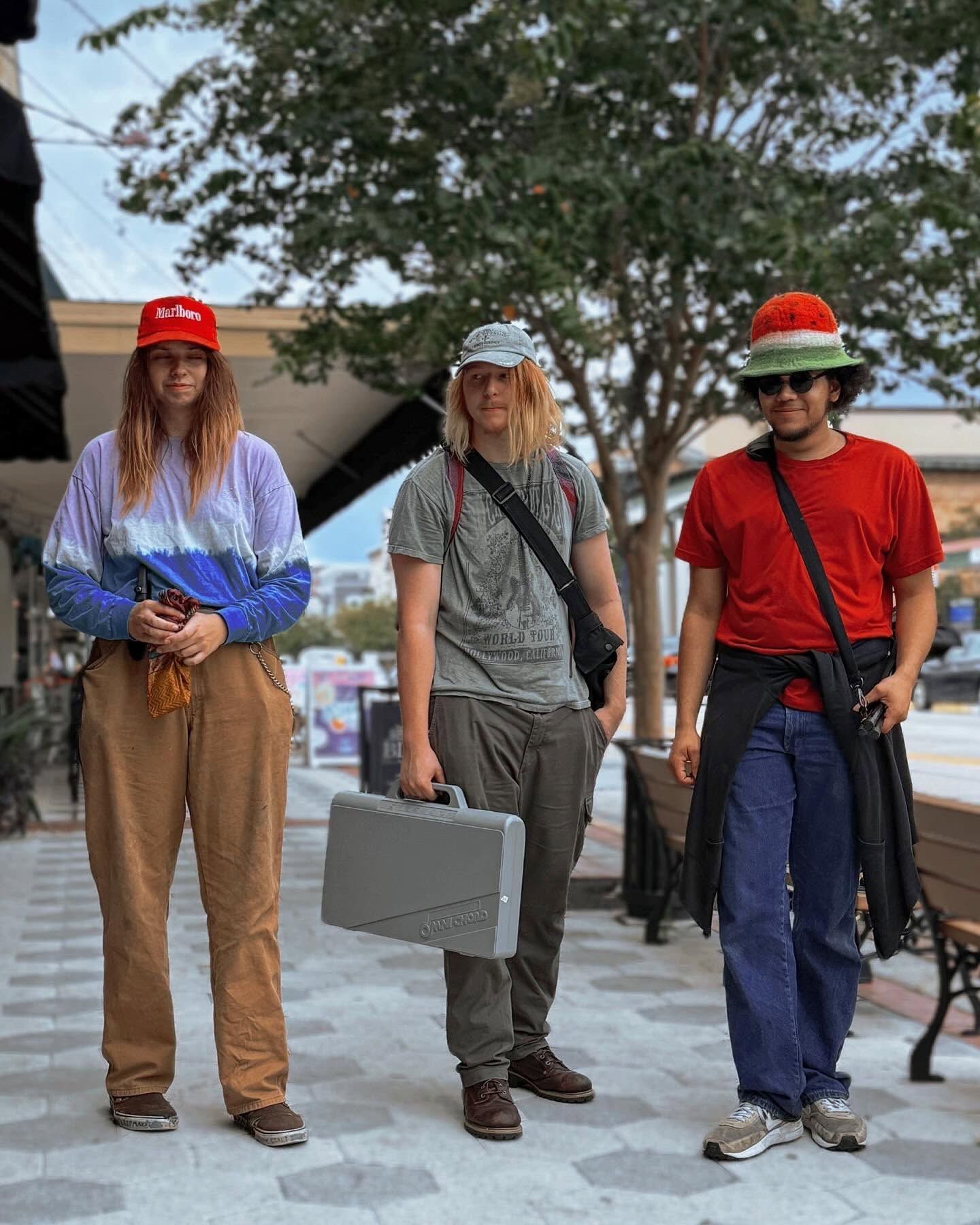 Three people standing outdoors, wearing red hats and holding a gray case in the center.