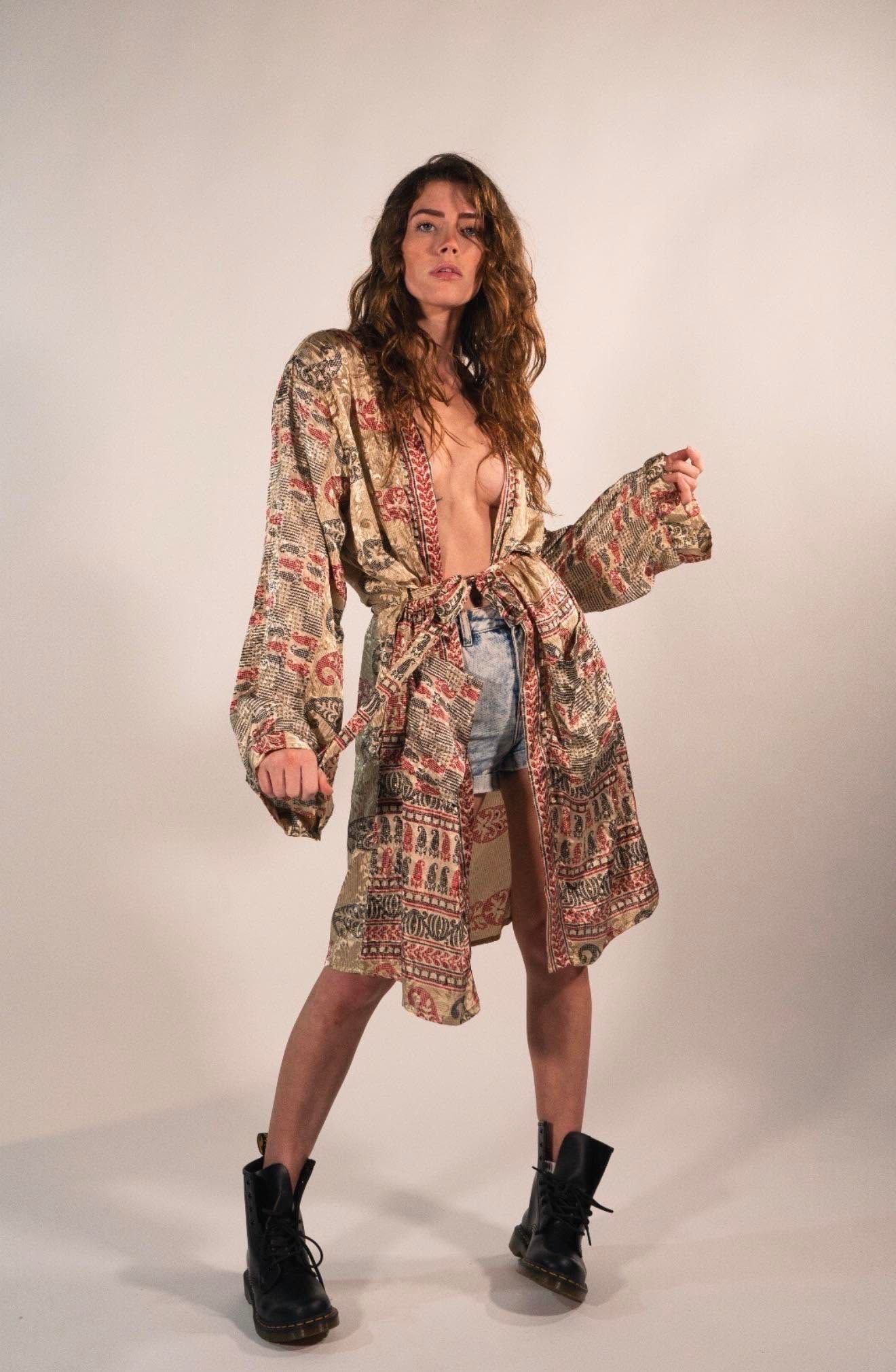 Woman posing in a patterned robe, blue jeans, and black boots against a plain studio backdrop.