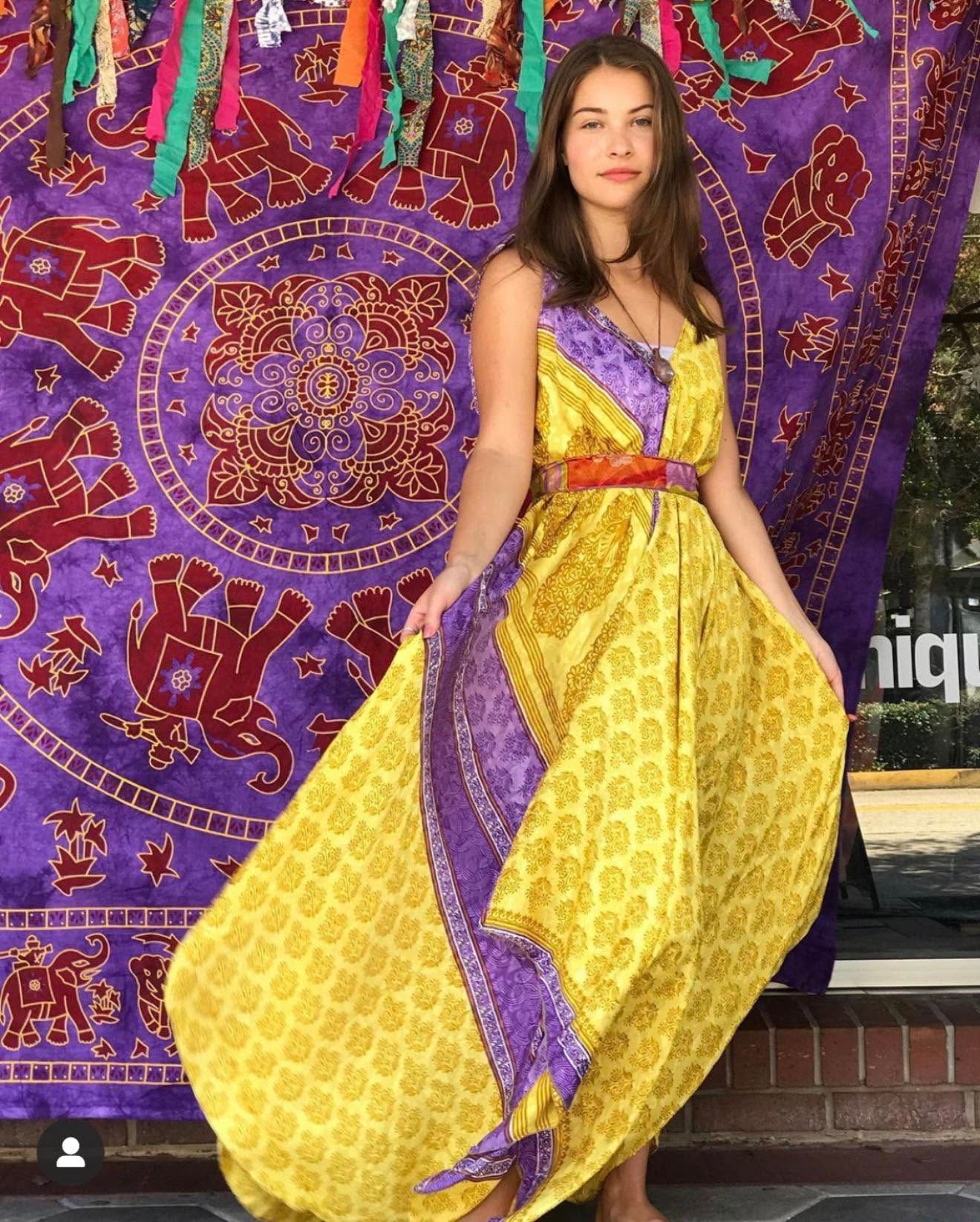 Woman in a yellow and purple dress posing in front of a colorful tapestry backdrop.