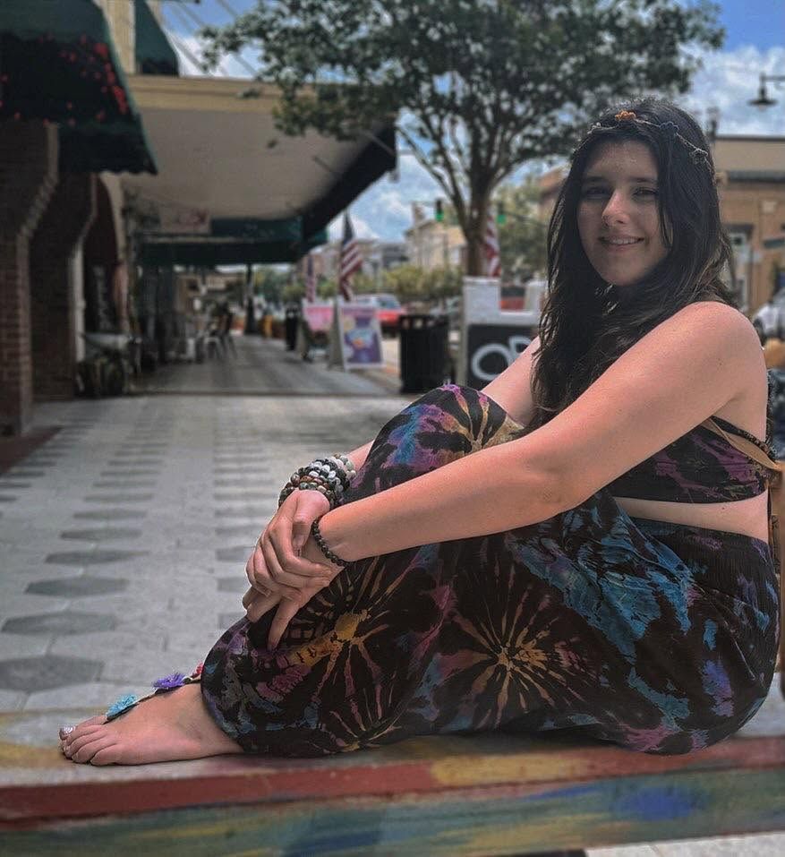 Person seated outdoors on a sidewalk, wearing a colorful patterned dress and smiling at the camera.