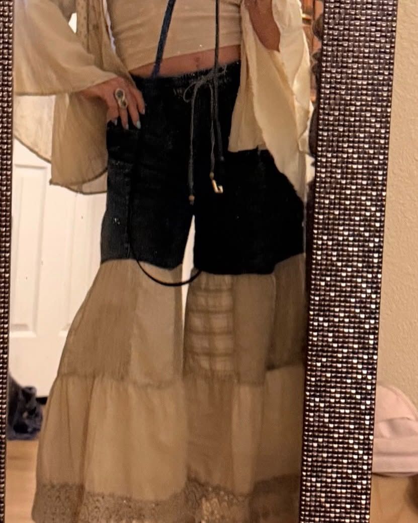 Person taking a mirror selfie in a cream jacket and black shorts over a beige tiered skirt.