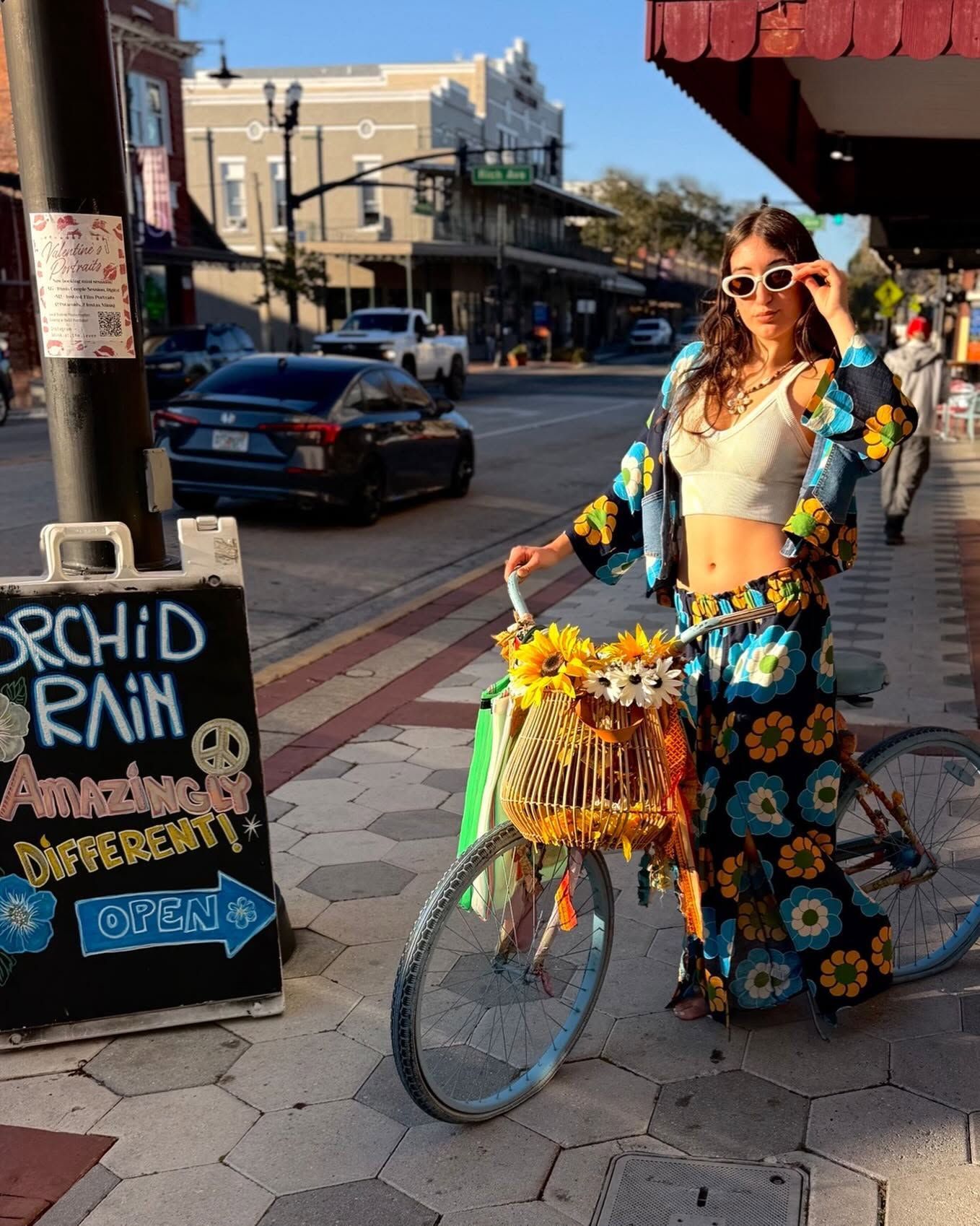 Woman posing beside a flower-decorated bicycle on a sunny street with storefronts and a sidewalk sign.