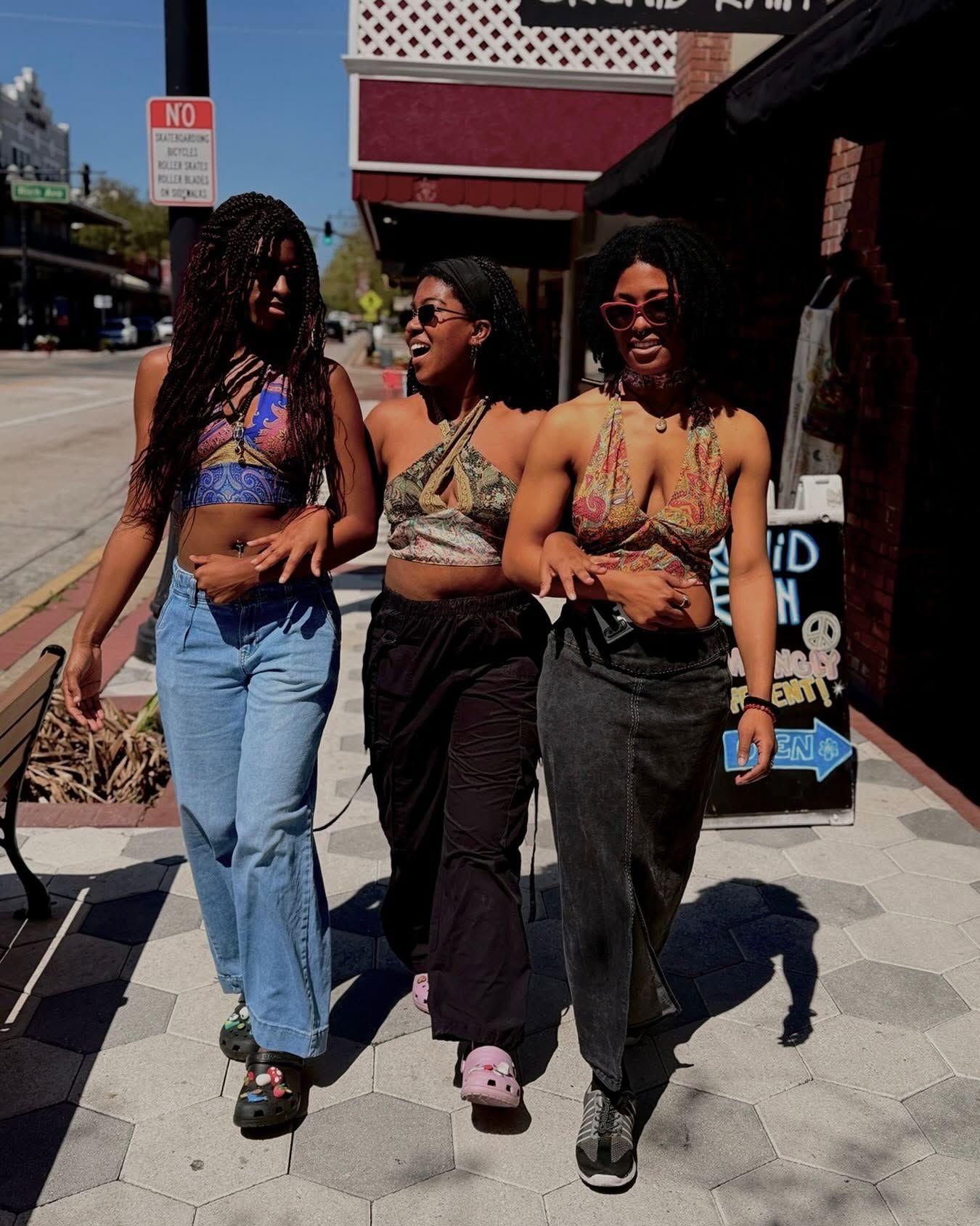Three friends walking arm-in-arm on a sunny city sidewalk, smiling and chatting.
