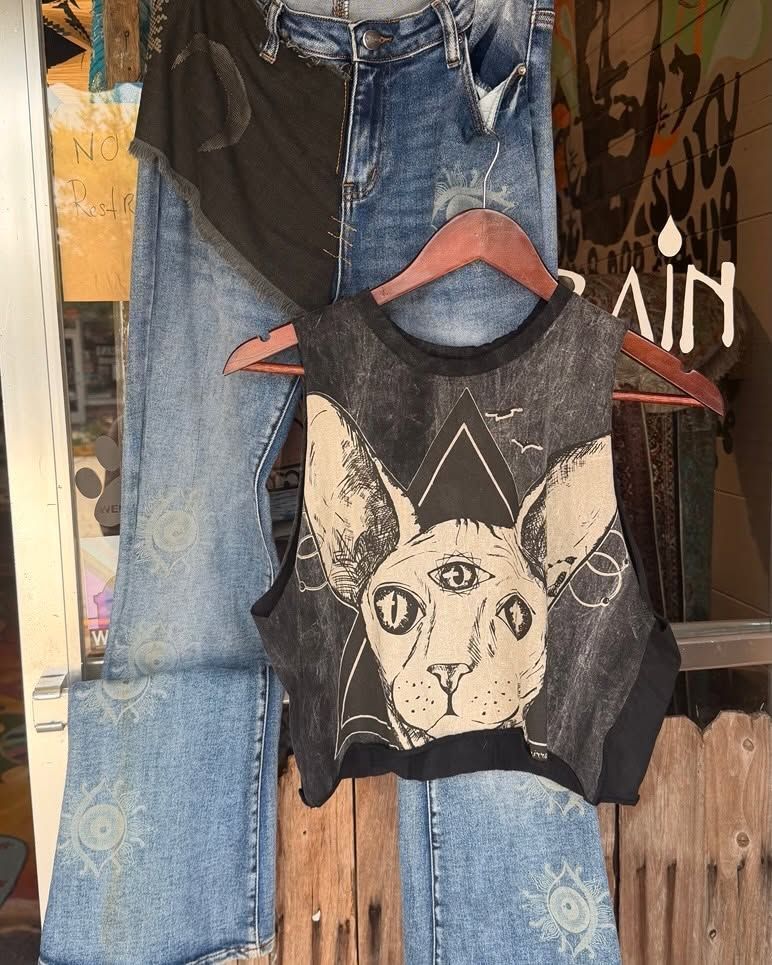 Black sleeveless cat-print shirt hanging with blue jeans on a rustic wooden fence.