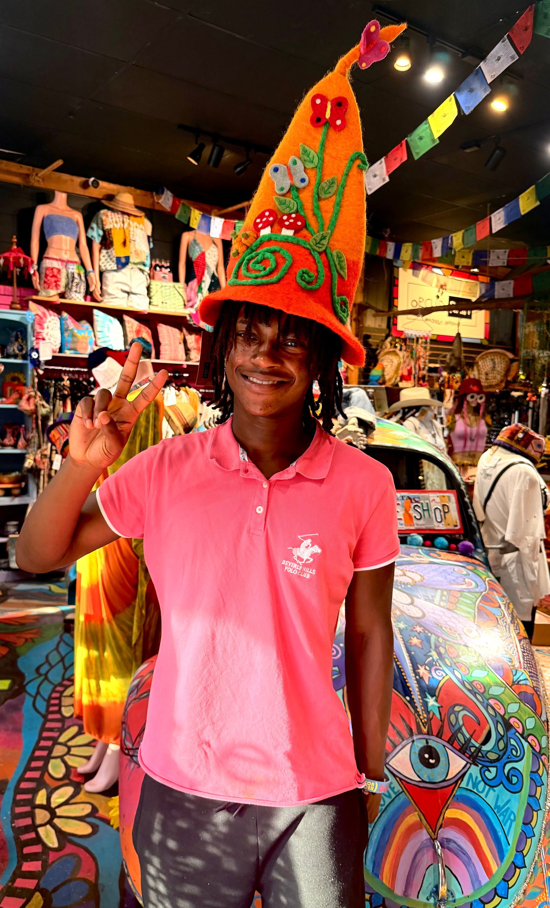 Person in a bright pink shirt smiling in a colorful souvenir shop, wearing a tall orange hat.