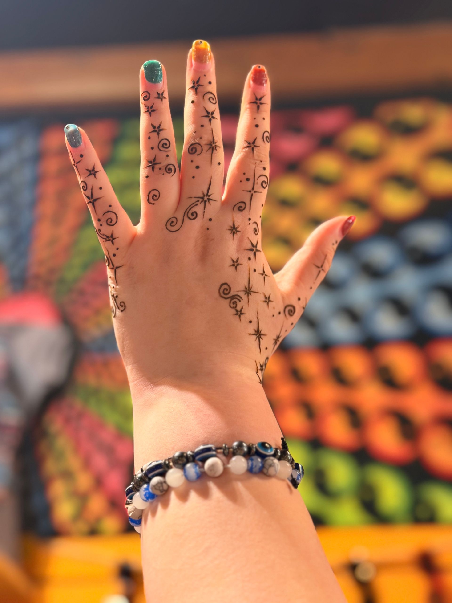 Hands with henna raised over a colorful patterned blanket, wearing a beaded bracelet.