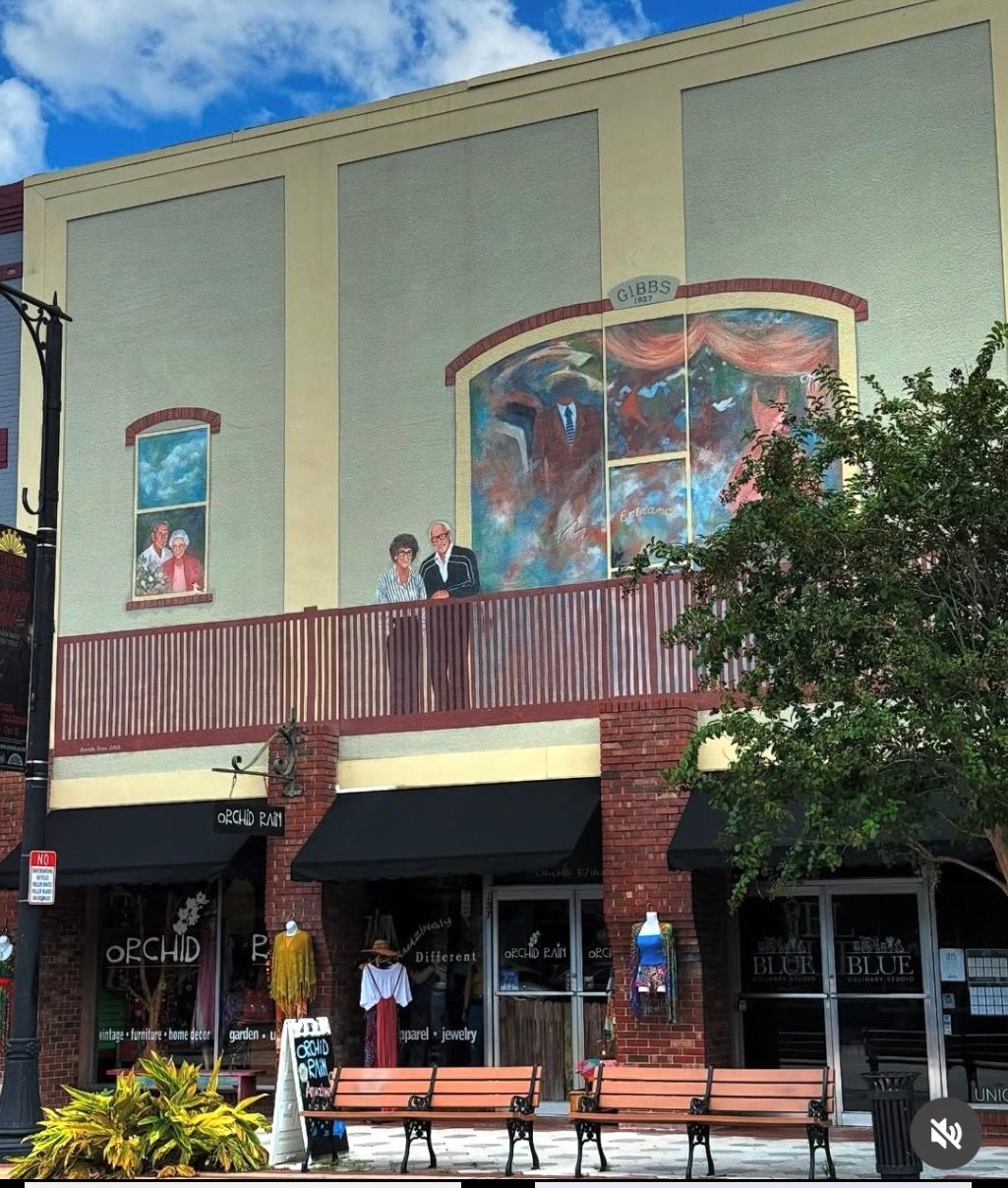 Street-front building with mural art, balcony, awnings, benches, and a tree in front