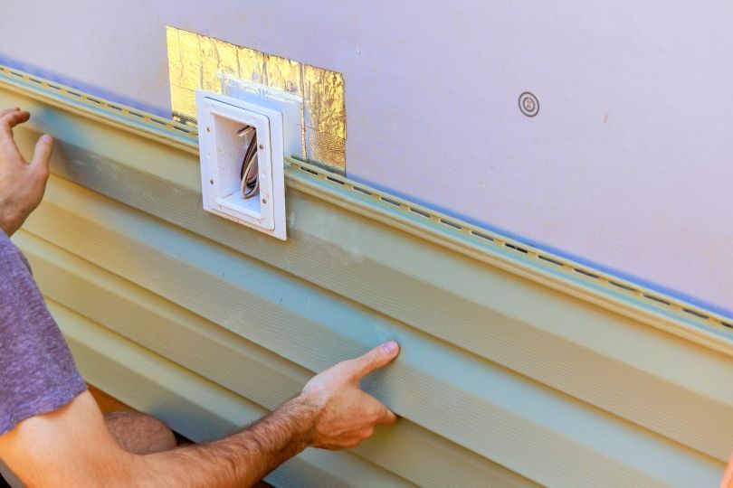 Person installing light green vinyl siding on a wall with a white electrical box.