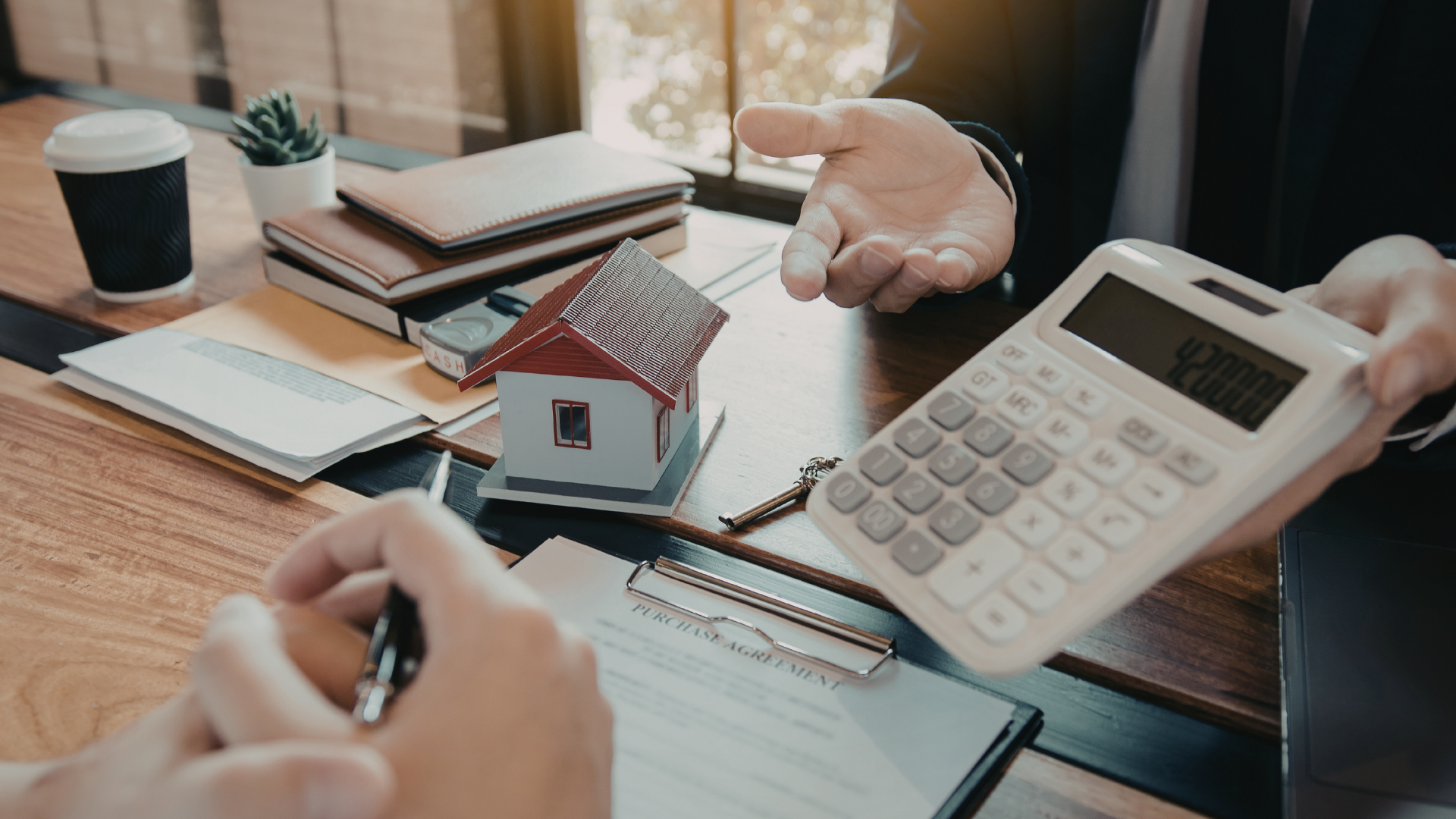 A person in a suit holds a calculator while another person reviews a document with a model house on a wooden desk.