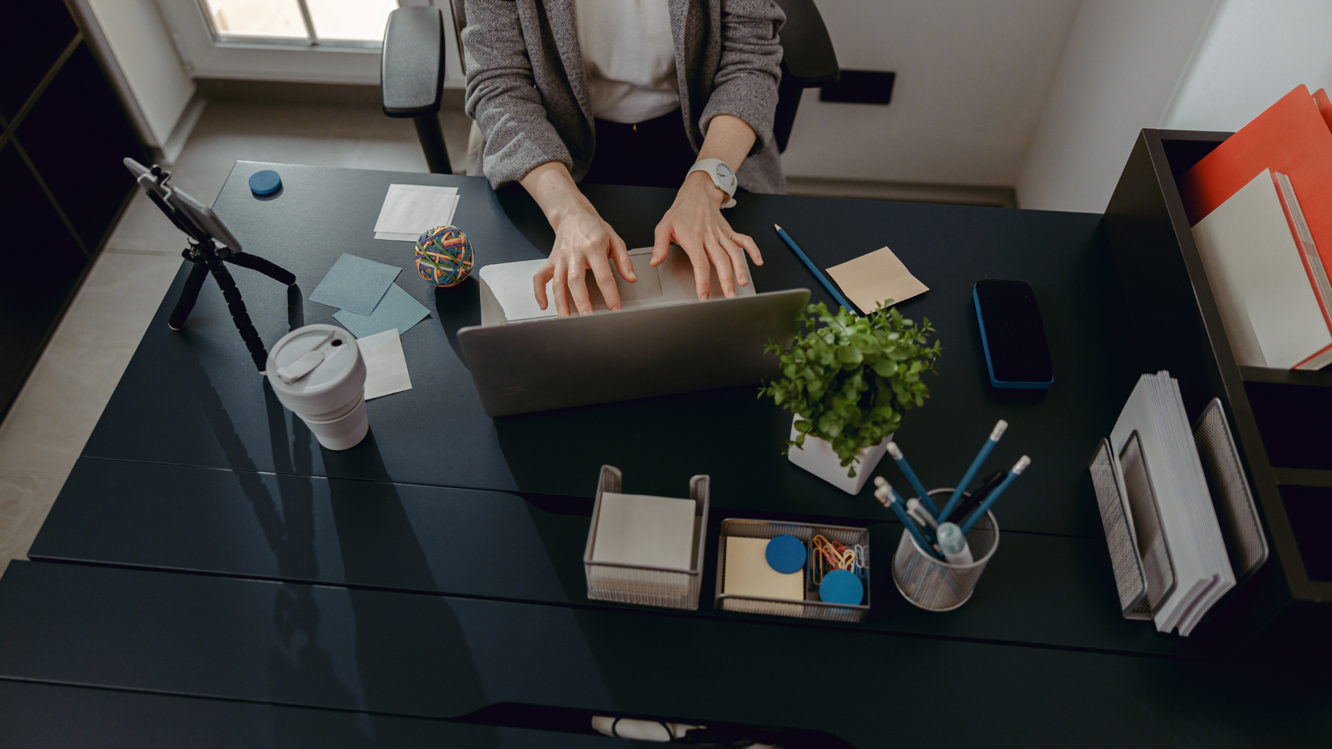 A high-angle view shows a person typing on a laptop at a dark desk, surrounded by a plant, stationery, and a mobile device.